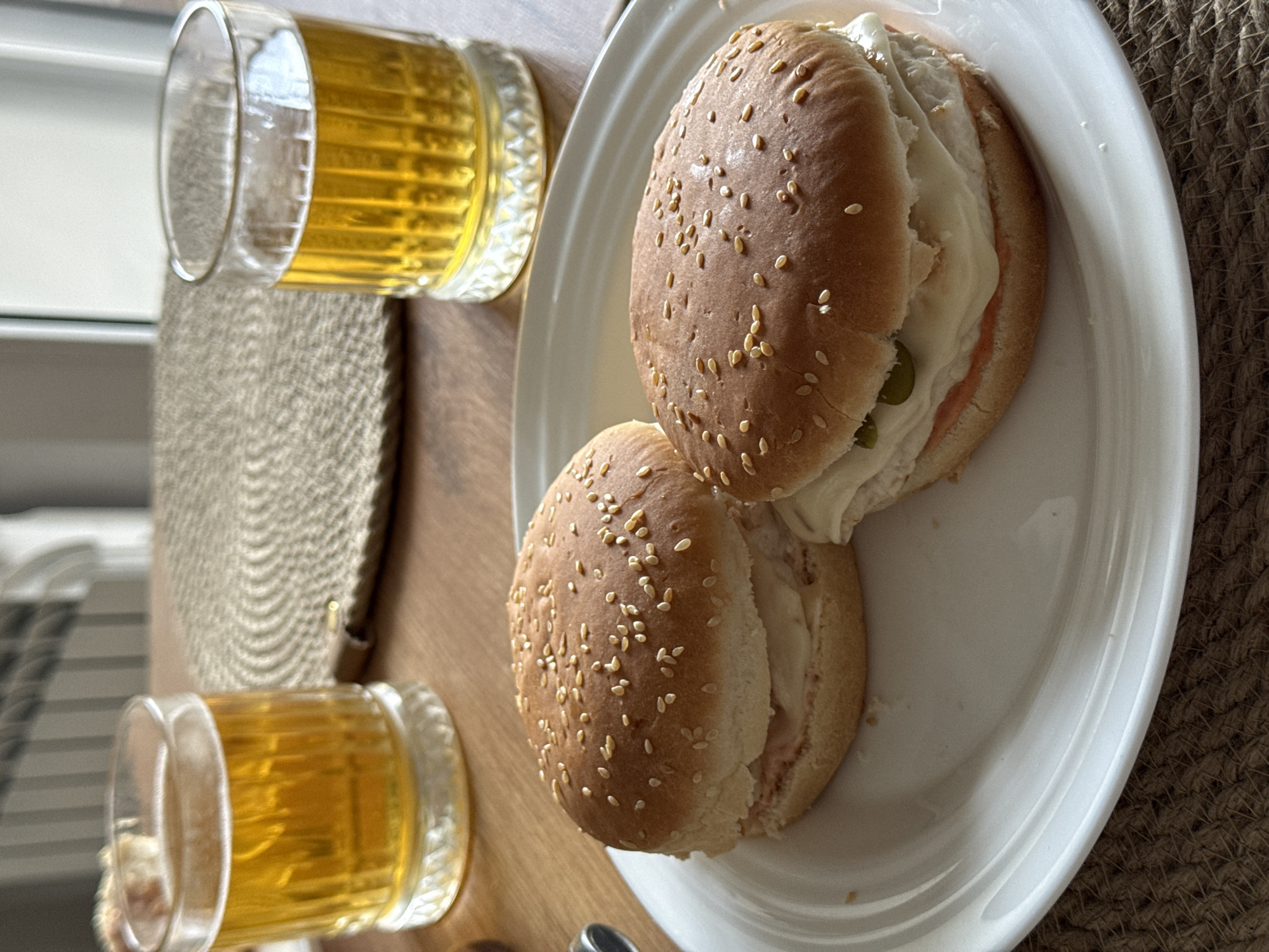 On a wooden table with a woven placemat, there is a white plate with two sandwiches and two glasses of amber-colored liquid. The sandwiches have sesame seed buns and appear to contain a filling with a white sauce, pink slices, and green vegetables. The glasses are cylindrical with a wide base and appear to be partially filled. The location is Новая Разводная, Russia.