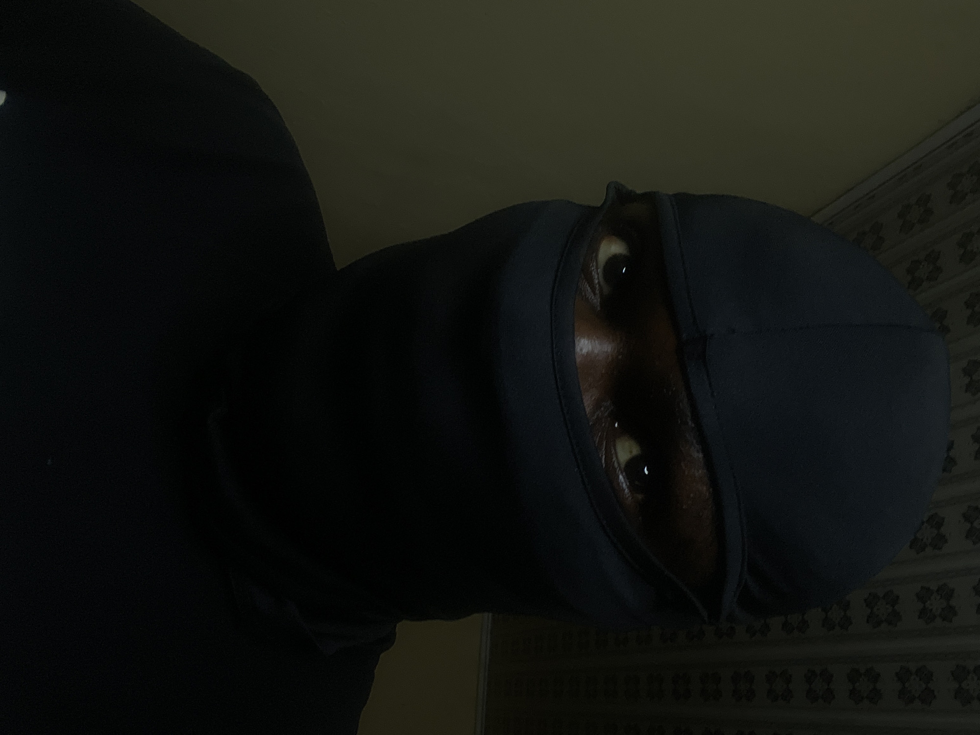 A low-angle, close-up shot shows a person wearing a dark-colored hoodie and what appears to be a durag over their head, obscuring most of their face except for their eyes. The person is looking down towards the camera. The background is out of focus, but appears to be a light-colored wall with a patterned design. There is no indication of the location of Jos, Nigeria in the image.