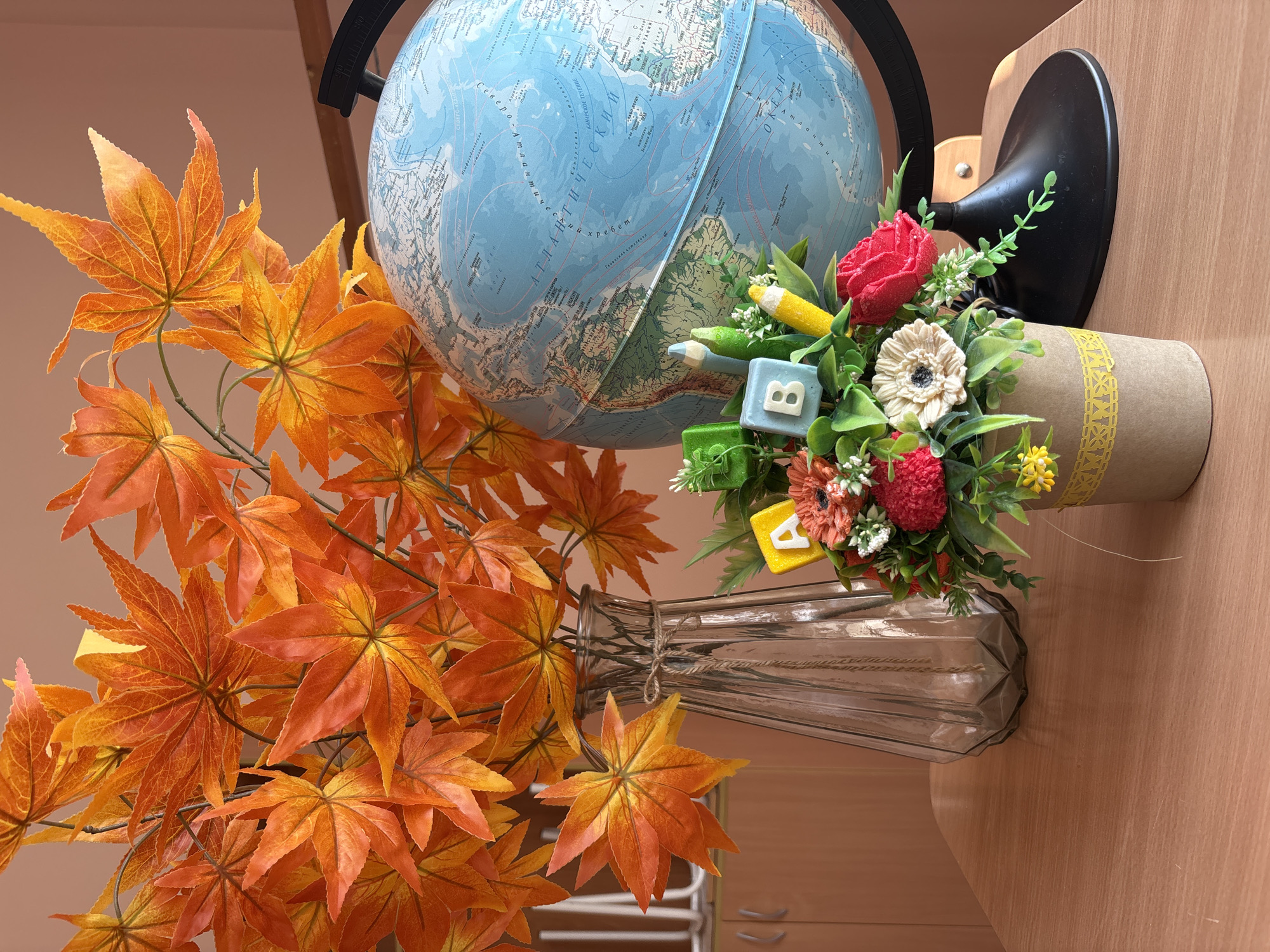 This is a still life image featuring a globe, a vase of artificial autumn leaves, and a small floral arrangement on a wooden desk. The globe is a prominent blue sphere with visible continents and oceans, resting on a black stand. To the left of the globe, a tall, clear glass vase contains a dense bouquet of bright orange and yellow artificial maple leaves. In front of the vase and to the right of the globe, a small pot holds a bouquet of artificial flowers in red, cream, and smaller yellow blooms, interspersed with green leaves. Several small, cube-shaped blocks with letters "A", "B", and "C" are placed amongst the flowers, along with two small, colorful pencils. The background shows a plain wall and a wooden cabinet. The scene is well-lit, with soft shadows indicating a light source from the right. The image does not contain any people or indicate a specific location within Korsovo, Russia.