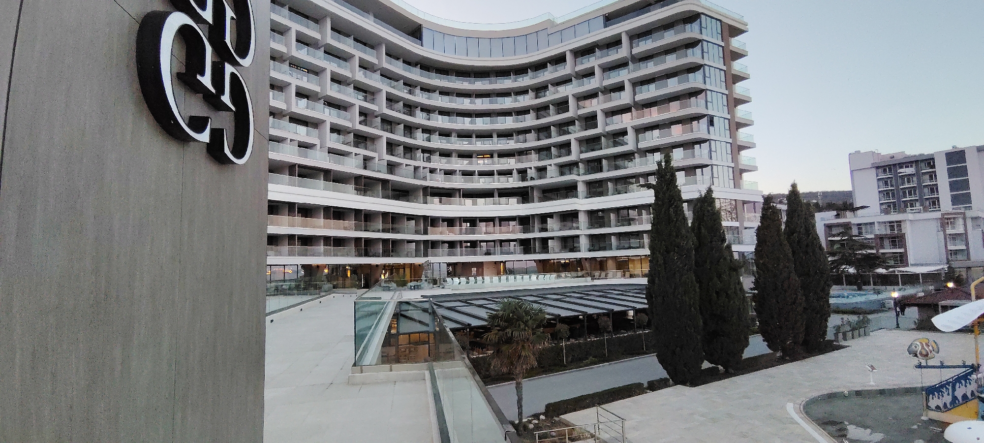 The image depicts a modern, multi-story curved building, likely a hotel or apartment complex, in Varna, Bulgaria. The building is white with glass balconies and windows. Adjacent to the building is a plaza with a covered outdoor dining area, landscaped with trees and plants. In the foreground, there's a partial view of a sign with a stylized emblem, possibly a logo. In the distance, another building can be seen, and to the right, there appears to be a playground area. The sky is overcast.