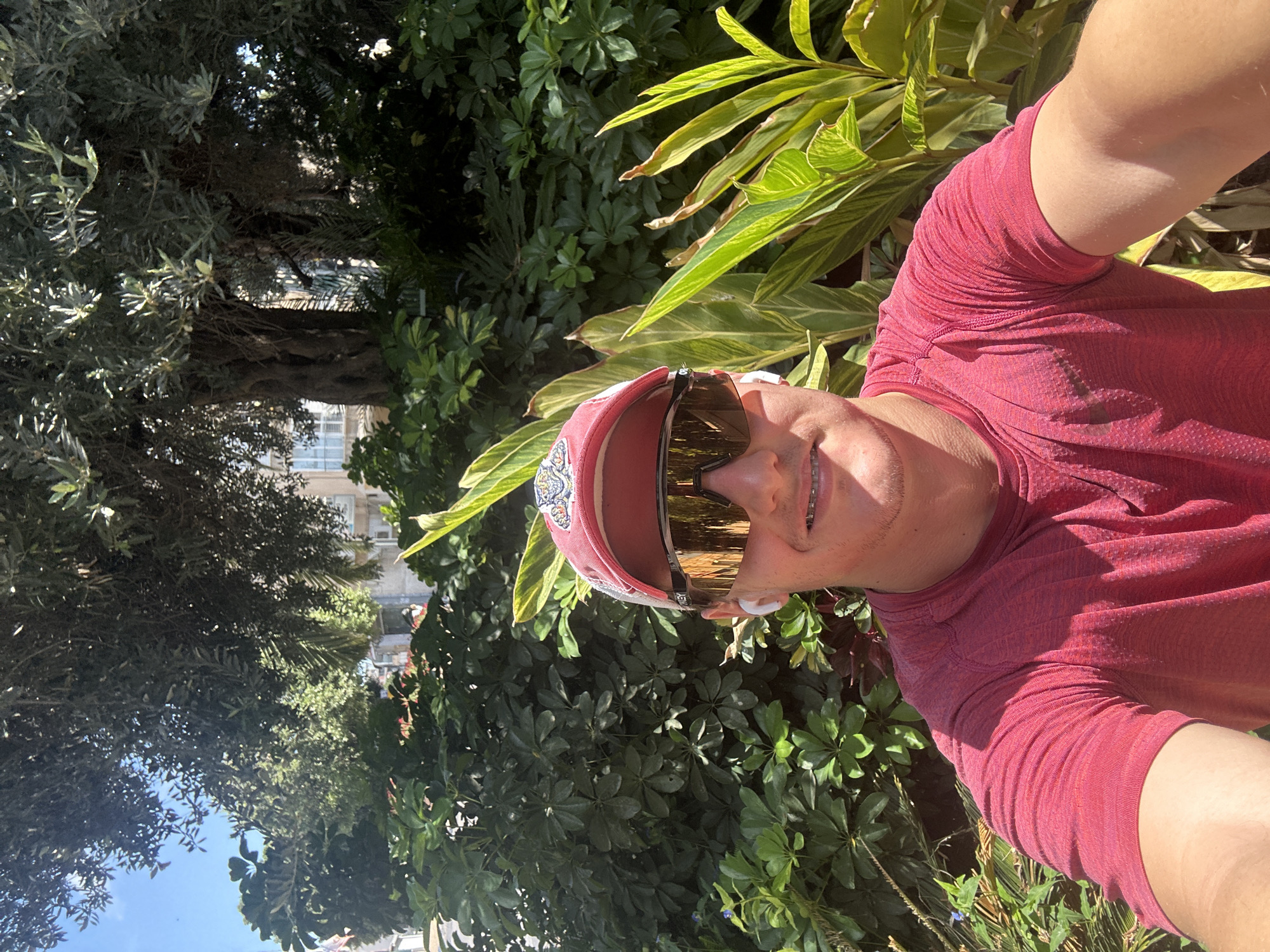 A selfie of a fair-skinned male wearing a red shirt, red and white baseball cap, gold Oakley sunglasses, and white earbuds stands among various green leafy plants. The location is Ramat Gan, Israel, discernible from the urban backdrop visible through the trees. The man is positioned in front of trees and plants, with a clear blue sky visible in the upper left corner.