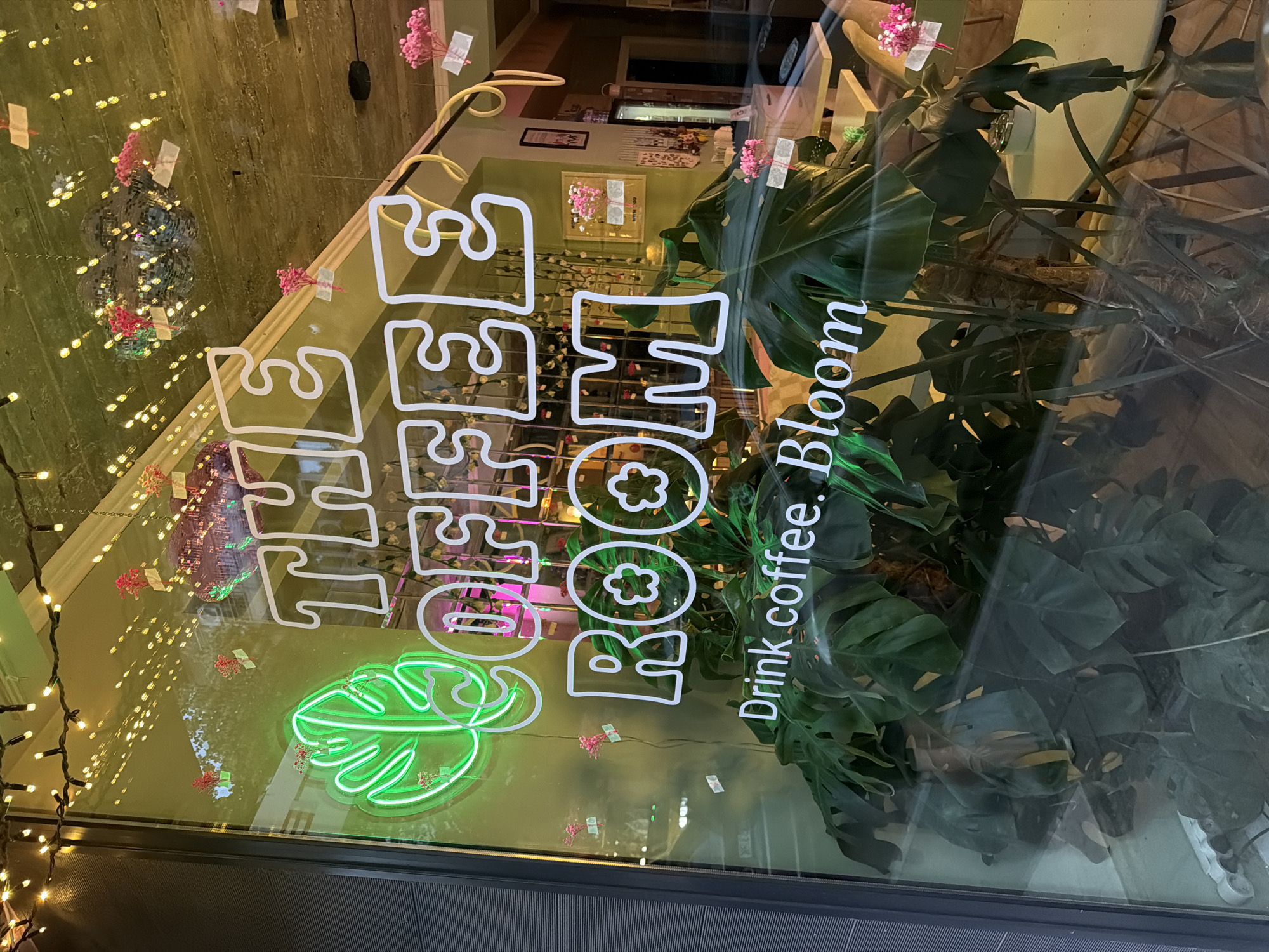 A neon sign in a shop window reads "THE COFFEE ROOM" above a green neon leaf emblem. Below the text, in white lettering, it says "Drink coffee. Bloom." Large tropical plants are visible inside the shop, obscuring much of the interior. Disco balls and string lights are also visible, reflecting in the window. The shop appears to be located in Burgas, Bulgaria.