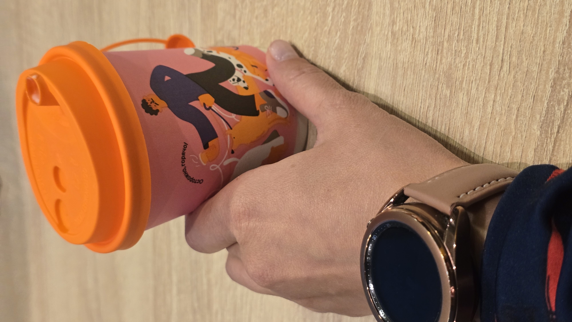 A close-up, eye-level shot shows a person's hand holding a pink disposable coffee cup with an orange lid. The cup has a graphic design of a person walking dogs. The person is wearing a rose gold smartwatch on their wrist. The background is a light-colored wooden surface, likely a table. The image does not contain any information that would allow for the determination of its location in Chaikovsky, Russia, or any other specific geographic location.
