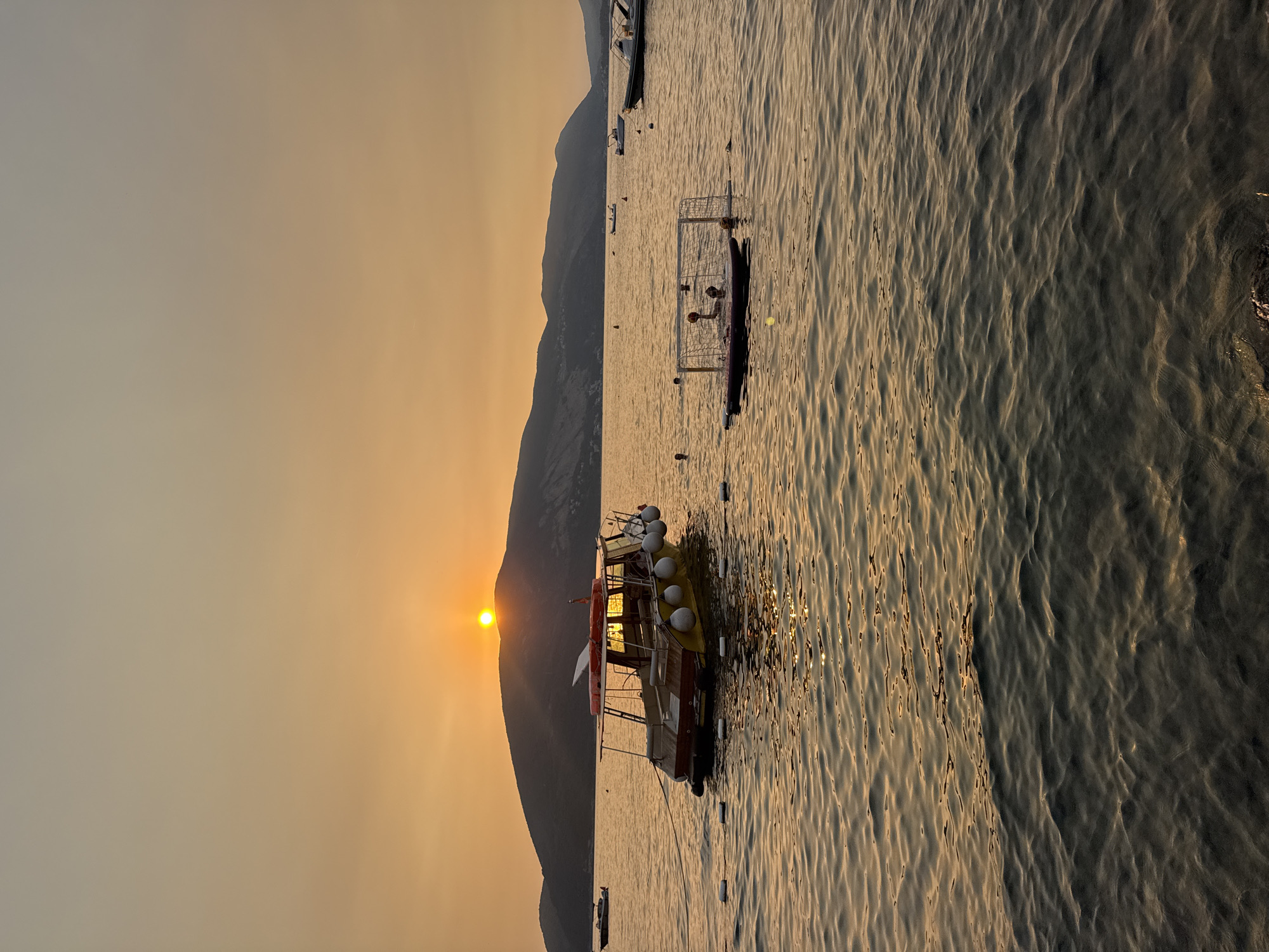 This is a vertical, eye-level, medium-shot photograph of a tranquil ocean scene at sunset. The sun, a bright orange orb, is partially obscured by a silhouetted mountain range in the distance. The sky is a soft gradient of orange and grey, indicative of the setting sun.
In the foreground, the water is calm with gentle ripples that reflect the warm glow of the sunset. Several objects are visible on the water. A yellow and red boat, with a canopy and several round white buoys attached to its side, is moored in the middle ground. To the right of the boat, a water polo net and several players are visible, indicating recreational activity. Further in the distance, more boats are scattered across the water. The overall mood is peaceful and serene.