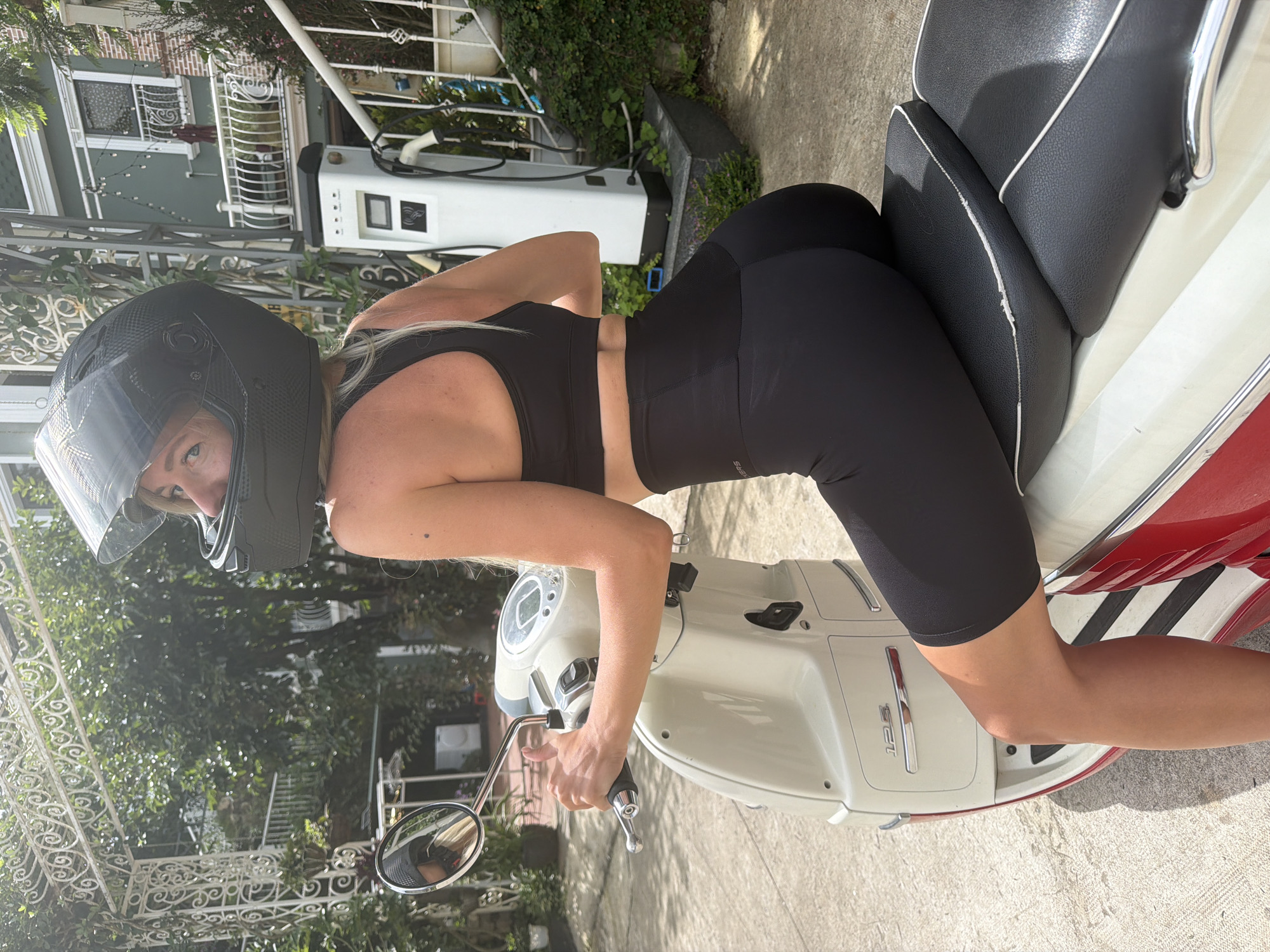 In the image, a woman wearing a black helmet and black athletic wear is posing on a cream and red scooter. She is positioned on the scooter, leaning forward with her hands on the handlebars. Her helmet covers most of her head, but her face is visible, and she appears to be looking at the camera. The scooter is parked on a paved surface, with a building and lush greenery in the background. An electric vehicle charging station is also visible in the background, suggesting a modern, potentially urban or suburban setting. The lighting indicates it is daytime, likely sunny, with shadows cast on the ground.
The image has been rotated vertically, so the scene appears sideways. The woman's athletic attire and helmet suggest she is either about to ride or has just finished riding the scooter. The overall mood of the image is casual and perhaps a bit playful. No visible text is apparent in the image itself, other than what might be emblems on the scooter or charging station, which are not clearly legible. The presence of the charging station could imply an eco-conscious or technologically advanced environment. The scene depicts a moment of leisure or preparation for an activity.
