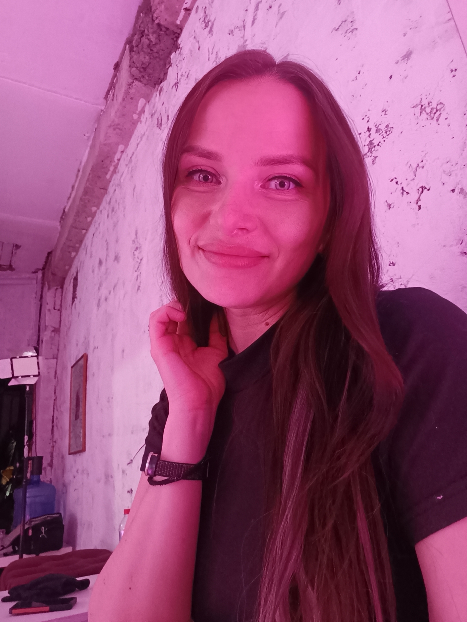 A young woman with long, straight brown hair is smiling and looking directly at the camera. She is wearing a black t-shirt and a black watch on her left wrist. Her left hand is raised near her neck. The background is a textured white wall, partially illuminated with pink light. In the lower left corner, there is a table with some objects including a water bottle and what appears to be a glove or small cloth. A light stand and other equipment can be seen blurred in the background. The location is indoors, in the city of Unknown, Unknown.