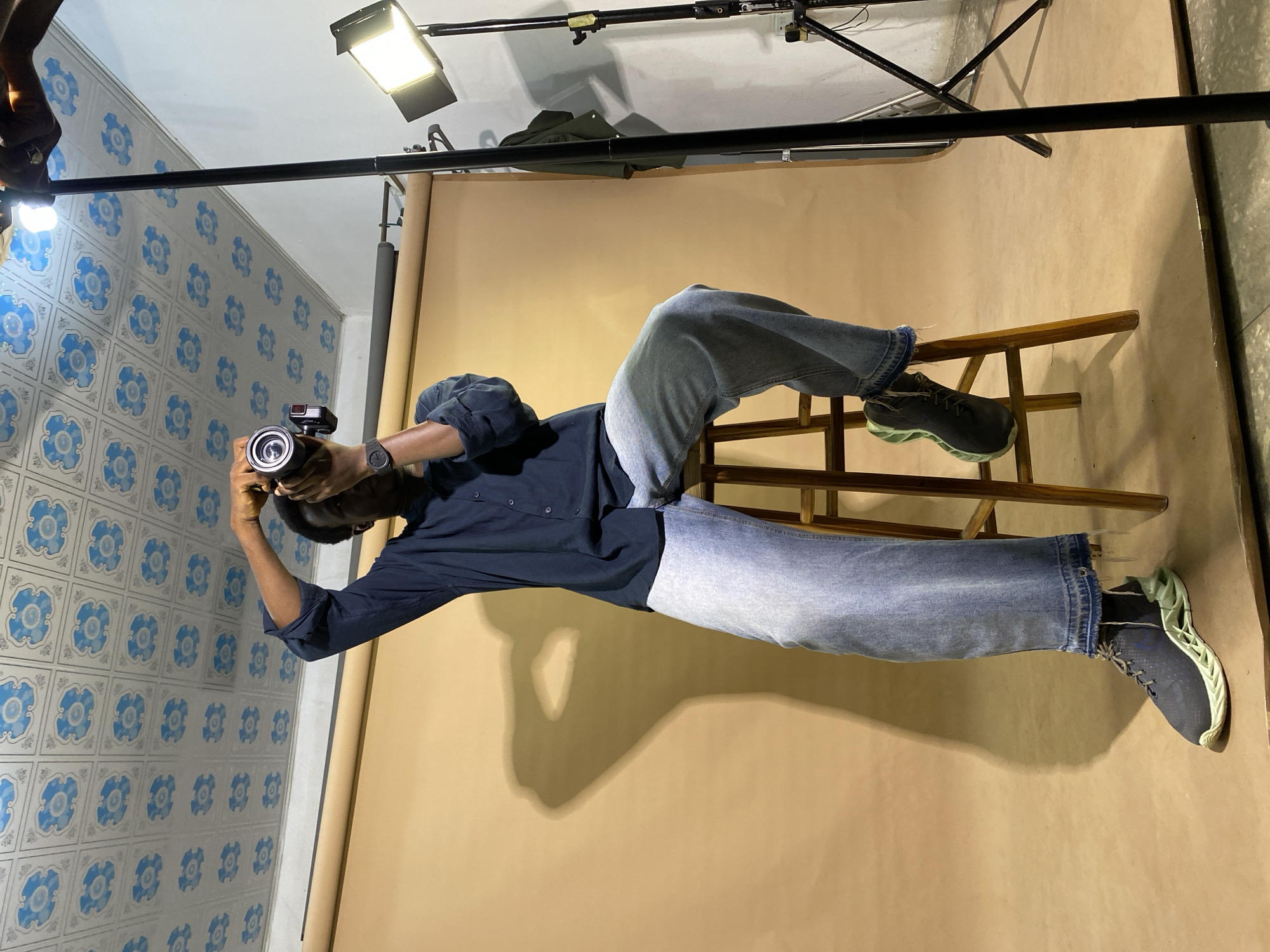 A dark-skinned man is seated on a wooden stool, holding a camera to his eye and seemingly taking a photograph. He is wearing a dark blue long-sleeved shirt and light-wash denim jeans. His right hand is raised to operate the camera, while his left arm is bent at the elbow. The man is positioned in front of a plain, tan-colored backdrop. To his right, a studio light is visible, mounted on a stand. The ceiling above features a patterned design of blue and white tiles. The scene appears to be an indoor photography studio. The man's shadow is cast prominently on the backdrop to his left. The ground surface is a mix of concrete and possibly a floor mat. The overall environment suggests a controlled setting for a photoshoot.