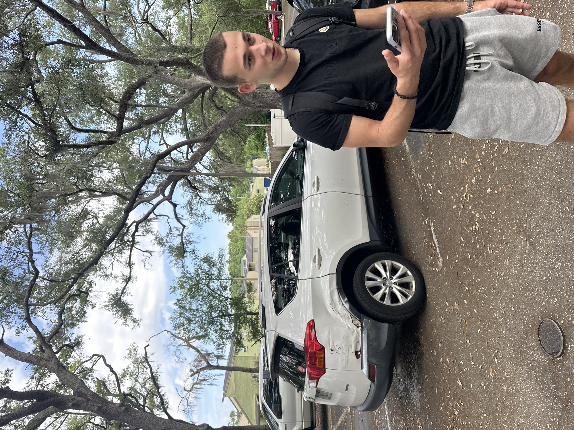 A young adult male is standing next to a white SUV with damage to the rear quarter panel, potentially in the city of University, United States of America. The man is holding a cell phone and wearing a black t-shirt, grey shorts, and a backpack. The SUV is parked on a paved surface near trees.