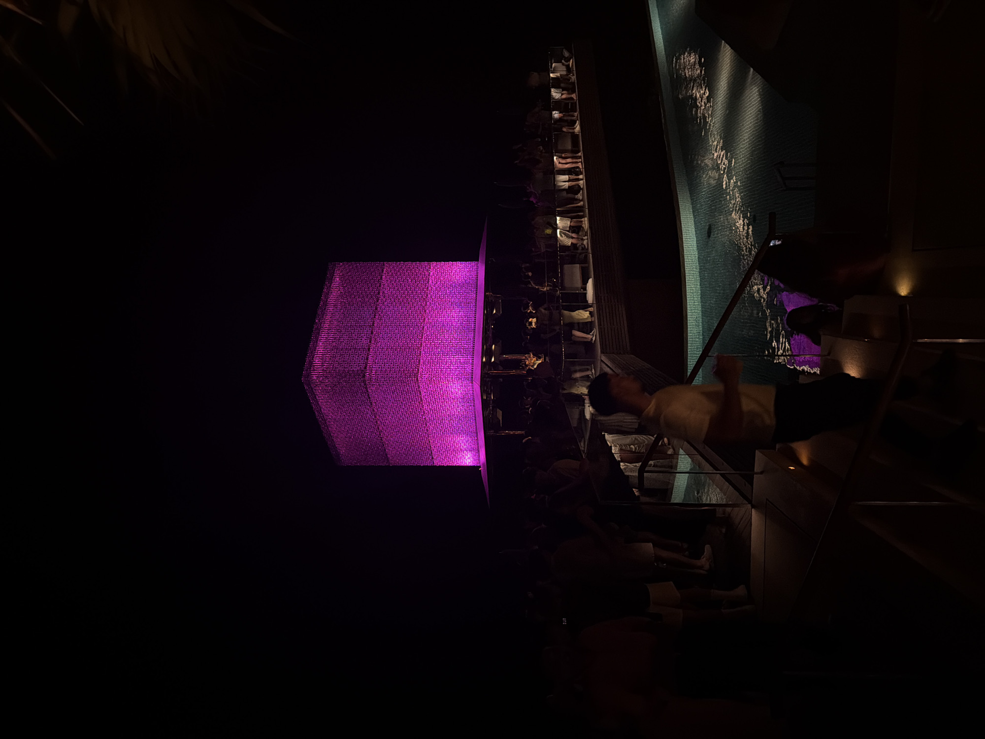 An exterior nighttime shot captures a vibrant outdoor venue with a striking cubic structure illuminated in bright pink. The cube, likely a building or stage element, sits atop a wider platform with a roof, where a crowd of people is gathered. Some individuals appear to be standing or socializing, while others are in motion. Below this elevated area, a swimming pool with textured tiles is visible, reflecting the ambient light and creating shimmering patterns on its surface. The overall scene suggests a lively atmosphere, possibly a party or event, at a location in Kuta Selatan, Indonesia, given the context of the provided crops. The foreground is dark, with a railing and a person walking down stairs, indicating a multi-level setting.