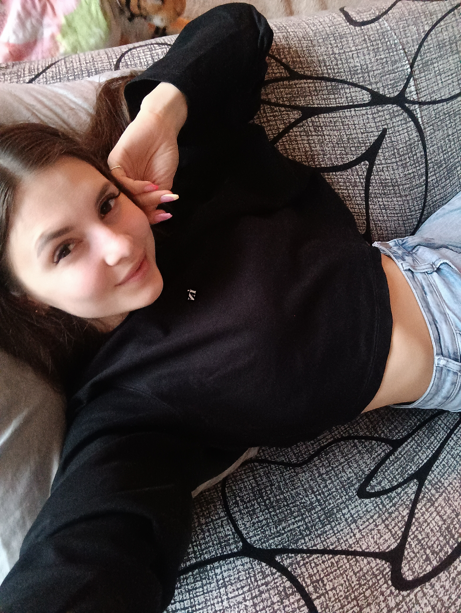 A young woman lies on a couch with a grey and black patterned fabric. She is wearing a black, long-sleeved cropped shirt and light blue jeans. Her left hand is near her face, showing pink and white nail polish. She is looking at the camera with a slight smile. The location is a residential setting and there is no evidence to suggest it is in the city of Chaikovsky, Russia.