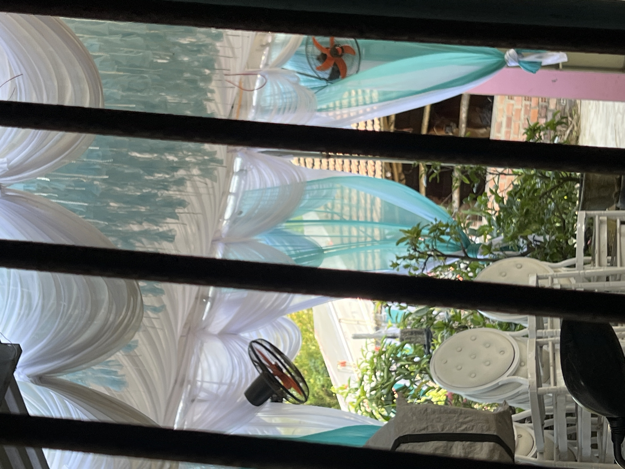 The image, observed through dark, horizontal slats that frame the view, depicts an outdoor or semi-outdoor event space under preparation. The scene is dominated by elegant fabric decorations, featuring white and light teal or mint green drapes intricately swaged and pleated overhead, creating a canopy effect.
Several small, portable fans with orange or red blades are visible within the decorated area, suggesting efforts to provide airflow in warm conditions. One fan is prominent in the upper right quadrant, and another is seen towards the lower left.
On the right side of the frame, several white chairs with round, tufted cushions are stacked, indicating ready-to-use seating for an event. Lush green foliage, including bushes and parts of trees, is visible throughout the background and interspersed with the decorations, confirming an outdoor setting. Distant background elements include a brick wall, some wooden structures, and a pink beam.
The bright, natural lighting suggests it is daytime. The setup, including the decorative fabrics, fans, and chairs, points to preparations for a formal gathering or celebration, likely a wedding or party, taking place in Bình An, Vietnam. No visible text is present in the image.