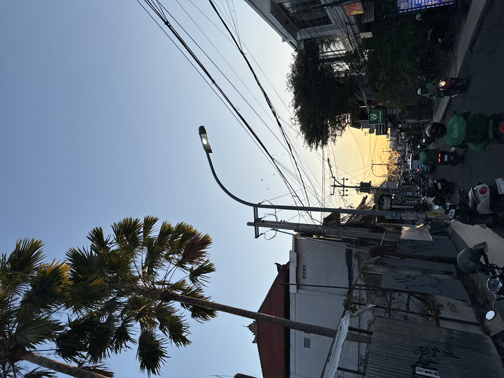 The image depicts a street scene in Kuta, Indonesia, during what appears to be late afternoon or early evening, with a clear, pale blue sky dominating the upper portion. Palm trees with prominent fronds are visible on the left, casting shadows. The architecture suggests a tropical location, with tiled roofs and buildings lining a street. The street itself is busy with motorbikes, some ridden by individuals wearing helmets. Several motorbikes are parked along the side of the road. One motorbike in the foreground has a license plate visible. There are also cars on the road, including a green vehicle. Numerous electrical wires are strung across the scene, supported by utility poles. Streetlights are present, with one illuminated. Signs for businesses, including "Franki Gardenia" and "MEGA MONEY CHANGER," are visible on the buildings. The overall impression is of a bustling urban environment in Indonesia at dusk.