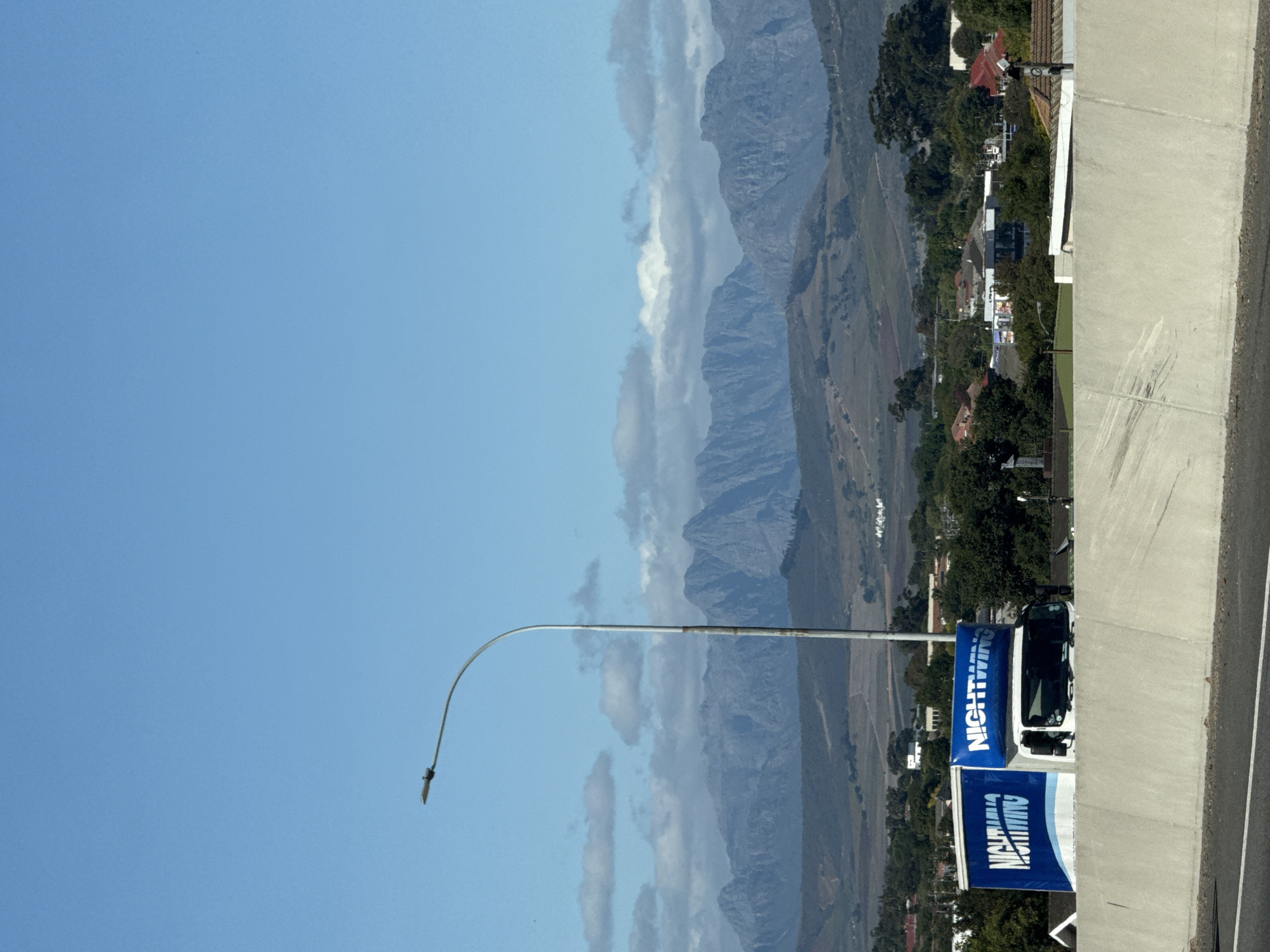 This image captures a wide, scenic view of a mountainous landscape, likely taken from an elevated vantage point such as an overpass. In the foreground, a light-colored concrete barrier or wall runs along the right side of the frame, with a segment of road visible beyond it. A tall, slender street lamp pole with a distinctive curved arm stands prominently on the left.
Partially visible behind the street lamp and the barrier is a white truck or van. Attached to the side of this vehicle, and also positioned lower in the frame, are two striking blue banners. Both banners prominently feature the word "NIGHTWING" in large, white capital letters. The lower banner also includes smaller, white text beneath "NIGHTWING," though it is less distinct.
The mid-ground reveals a sprawling valley characterized by fields and natural vegetation, appearing in shades of green and brown. Numerous houses and buildings, some with red roofs and others with grey, are scattered across this area, suggesting a residential or rural settlement.
Dominating the background is a majestic mountain range, stretching across the entire width of the scene. The mountains display rugged peaks and slopes in tones of blue and grey, indicating distance. Patches of white and grey clouds are interspersed among and above the mountain peaks, contrasting with the expansive, bright blue sky. The clear sky and strong illumination suggest it is daytime, likely late morning or early afternoon, with fine weather. The scene conveys a sense of peaceful natural beauty. No specific human activity or individuals are visible. The location context is Bellville, South Africa.