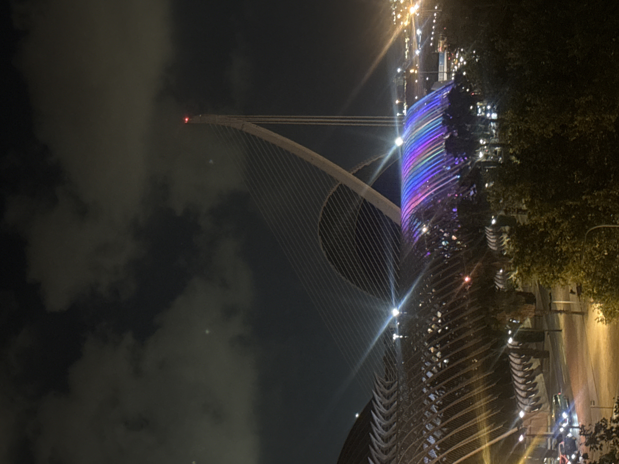 The image depicts the City of Arts and Sciences in Valencia, Spain at night. The L'Umbracle, a landscaped promenade with arcades, is illuminated with colorful lights, displaying a rainbow effect. To the right of L'Umbracle, the Assut de'l'Or Bridge is visible, its tall pylon topped with a red light. The sky is dark with scattered clouds. In the foreground, trees and a street with cars are present. The overall scene conveys a vibrant and modern urban nightscape.