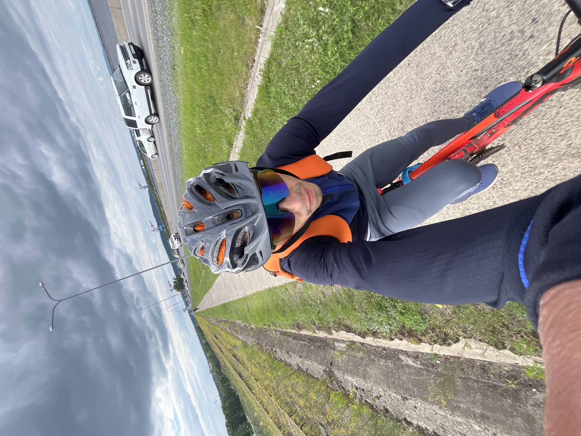 A man is riding a red bicycle on a paved path next to a grassy area with railroad tracks. He is wearing a helmet, sunglasses, a dark blue long-sleeved shirt, gray pants, and blue shoes. He has an orange backpack. The path appears to be in Chaikovsky, Russia, as there are streetlights and a highway with cars visible in the background. The sky is cloudy, suggesting overcast weather. The scene is captured from a first-person perspective, indicating the man is holding a camera or phone.