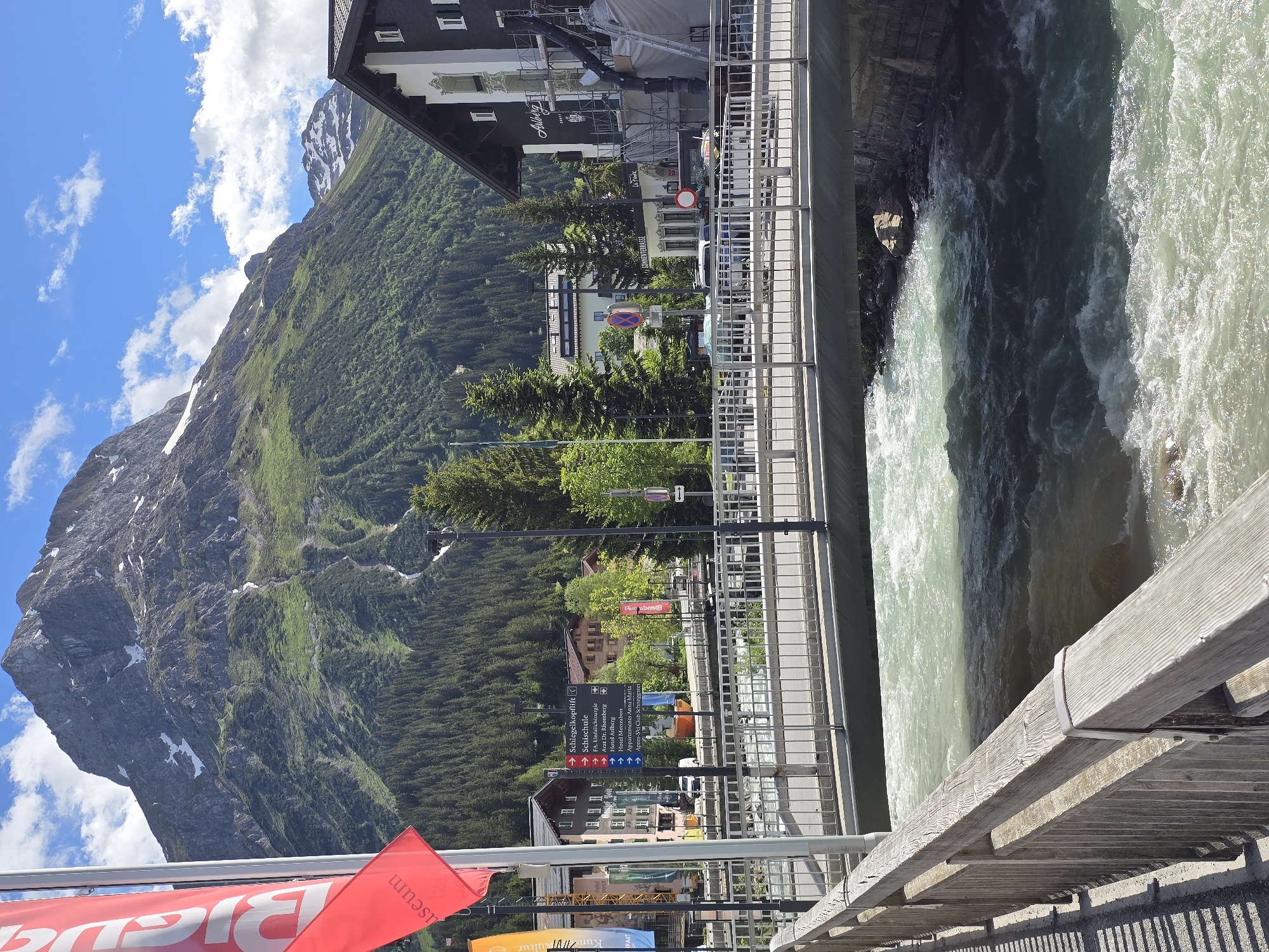 The image depicts a bright, sunny day in Lech, Austria, showcasing a vibrant mountain landscape and a bustling village scene. In the foreground, a wooden railing borders a footpath alongside a fast-flowing, turquoise river. The river, churned with white rapids, passes under a modern metal bridge. Beyond the bridge, a street lined with buildings, flags, and signage indicates the presence of a village center. The buildings display traditional alpine architecture with wooden balconies and dark-tiled roofs. Prominent among them is a building with "Alberg" signage. A large, imposing mountain dominates the background, its slopes covered in dark green coniferous forests, with patches of snow visible near its rocky, grey peak. The sky is a brilliant blue, dotted with fluffy white clouds. Several signs are visible, including one with directions to "Schlegelkopfift," "Schischule," and various hotels and apartments. Red and orange flags with white lettering are also present, adding pops of color to the scene. The overall impression is one of a scenic and active mountain resort town.