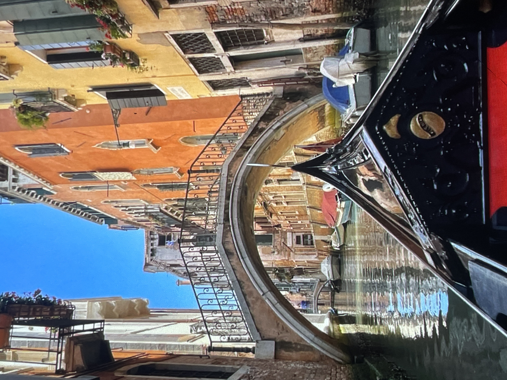 The image shows a canal in Venice, Italy, not Harmanli, Bulgaria. The scene is viewed from the perspective of a gondola. In the foreground, the ornate black and gold decoration of the gondola is visible. The canal is lined with colorful buildings, predominantly orange and yellow, with many windows and balconies. A pedestrian bridge arches over the canal in the mid-ground, with people visible on it. Several smaller boats are moored along the canal. The water reflects the buildings and the blue sky.