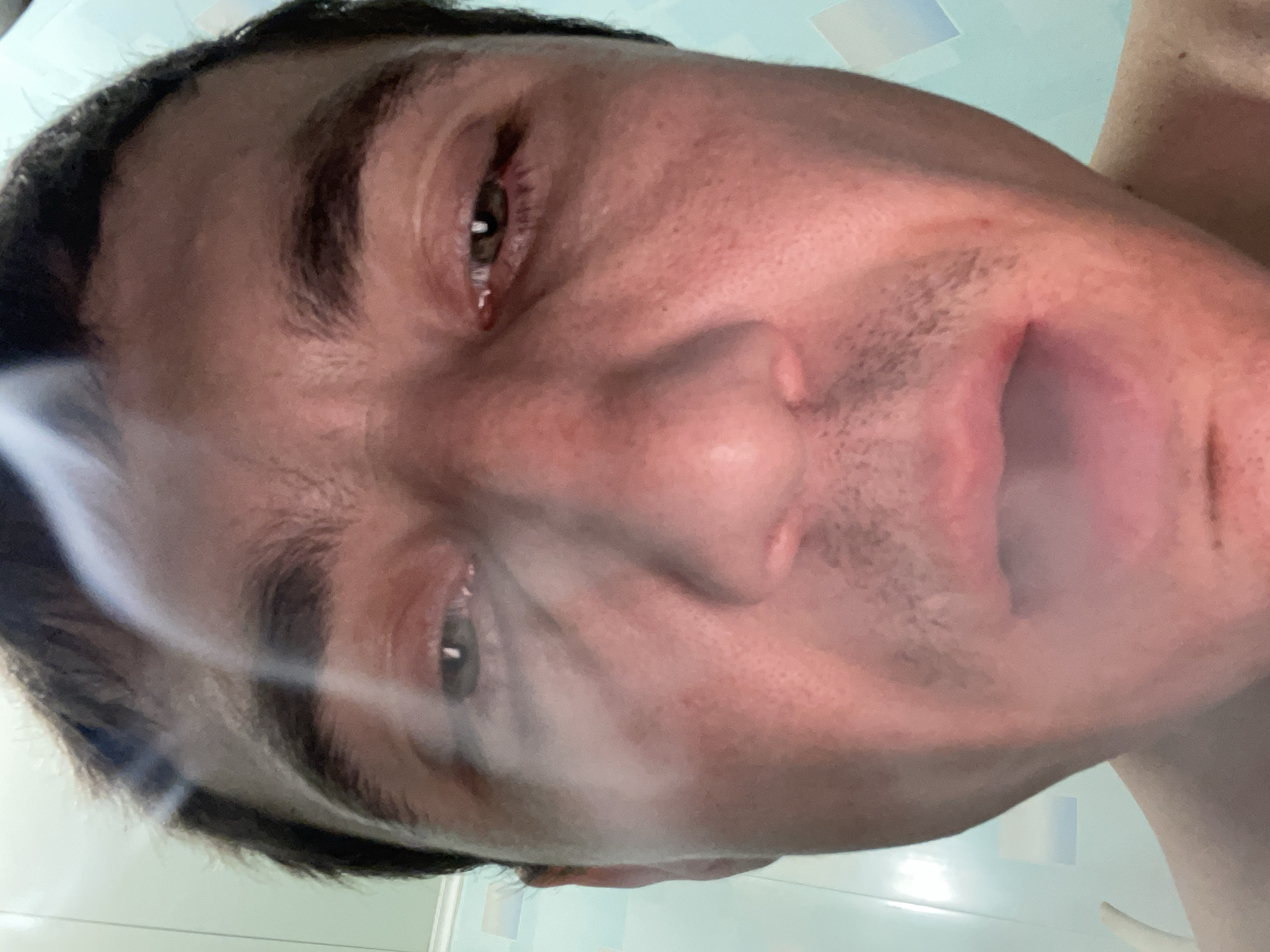 This is a close-up, low-angle, selfie-style photograph of a person's face, taken indoors. The subject, appearing to be male with short, dark hair and fair skin, is looking directly at the camera. Their eyes are open but appear somewhat tired or strained, with one eye slightly red. A thin plume of white smoke drifts across the upper portion of the face, obscuring parts of the forehead and eyes. The mouth is slightly open, revealing the lower teeth and tongue. The skin shows some texture and subtle blemishes. The background is out of focus but appears to be a bathroom wall with a light blue and white tiled pattern. The lighting is harsh, creating strong highlights and shadows on the face. There is no indication of the city of Laishevo, Russia, within the image itself.