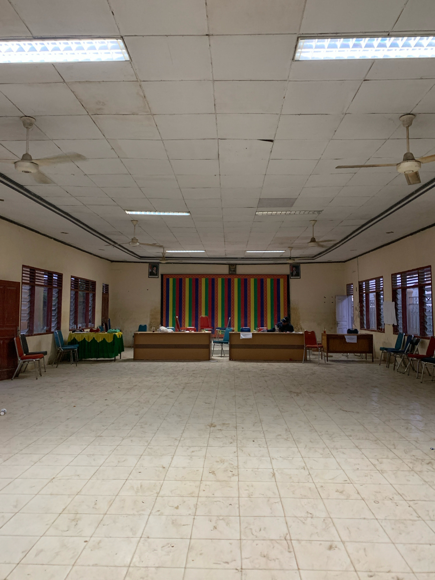 The image depicts an interior view of what appears to be a meeting room or hall, likely in Pantegurah, Indonesia, given the context. The room is rectangular with a tiled floor and a dropped ceiling with fluorescent lighting and ceiling fans. Along the walls are windows with slatted coverings.
In the foreground, the tiled floor shows signs of wear and is not perfectly clean, with some scattered debris.
Towards the rear of the room, there are two long tables arranged perpendicular to the back wall. The table on the left is draped with a green cloth and a yellow tablecloth. On this table, several chairs are visible, including blue and red chairs. The table on the right is a wooden desk with chairs in front of it, including red and blue ones.
The back wall features a colorful, striped banner or tapestry hanging centrally, with portraits of individuals framed on either side of it. Doors are visible on the side walls. There are no people clearly depicted in the image, though the arrangement of furniture suggests a space for gatherings or formal events. The overall impression is of a functional but somewhat dated community space.
