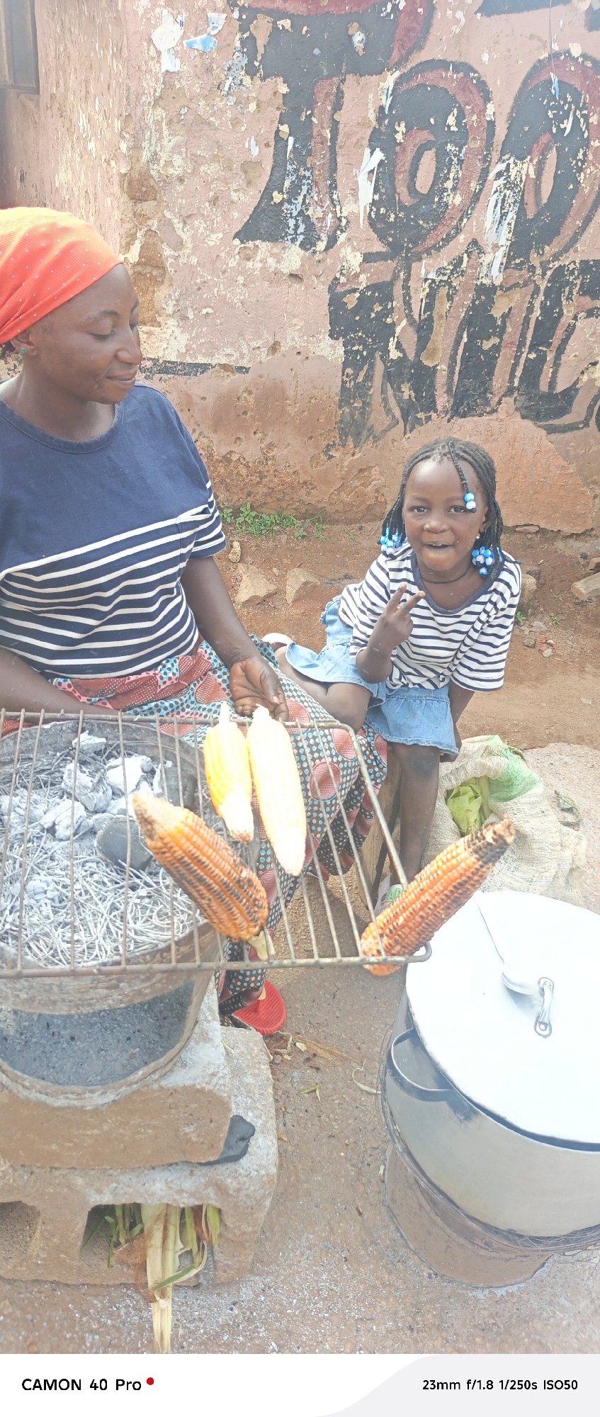 This media file captures a candid moment with two individuals, an adult and a child, at an outdoor food stall. The adult, a woman wearing an orange headscarf and a striped blue and white shirt, is seated and appears to be attending to a grill. The child, a young girl with braided hair adorned with blue beads, is seated next to her, facing the camera and making a peace sign with her fingers. She is wearing a similar striped shirt and a blue skirt.
The scene is set outdoors with a textured, worn wall in the background, featuring what appears to be graffiti or faded paint. In the foreground, a metal grill is positioned over hot coals, with several ears of corn cooking on it. One ear of corn is already quite browned, indicating it's nearly ready. To the right, a large pot with a lid is visible, suggesting preparation of other food items. A sack of what looks like produce or supplies rests behind the child. The ground is dirt and small stones.
The lighting suggests it is daytime, possibly morning or afternoon, with natural light illuminating the scene. The overall mood appears relaxed and ordinary, depicting a moment of daily life and commerce. The focus is on the people and the food preparation.
Visible text at the bottom of the image includes "CAMON 40 Pro" and "23mm f/1.8 1/250s ISO50", which are likely camera specifications. The provided location context is Barikin Gidar Kanat, Nigeria.
