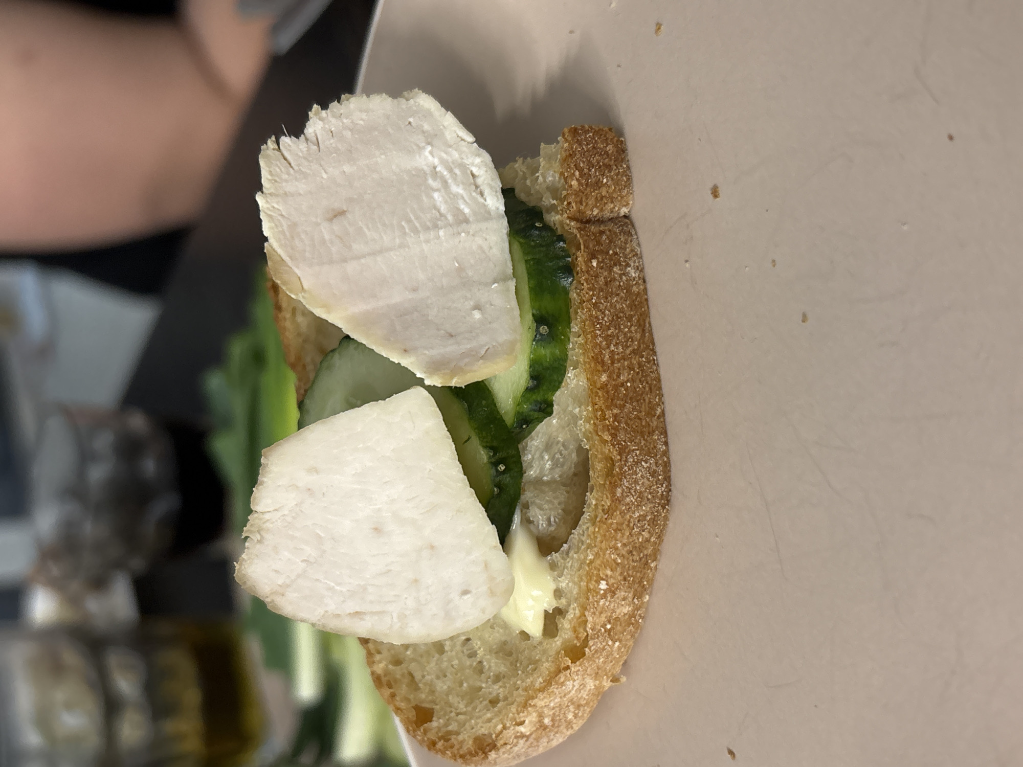 A close-up shot of a partially constructed sandwich is positioned on a light-colored, possibly off-white, surface. The sandwich consists of a piece of bread with a coarse crust, upon which is arranged sliced cucumber, lettuce, and two slices of what appears to be cooked poultry, possibly turkey or chicken. There is a pale-colored spread, likely mayonnaise or aioli, visible on the bread. The background is blurred, but a dark object is visible to the left, possibly a jar or bottle. Part of a person, specifically a shoulder and arm, is also visible in the upper left of the image. The location is not evident from this image.