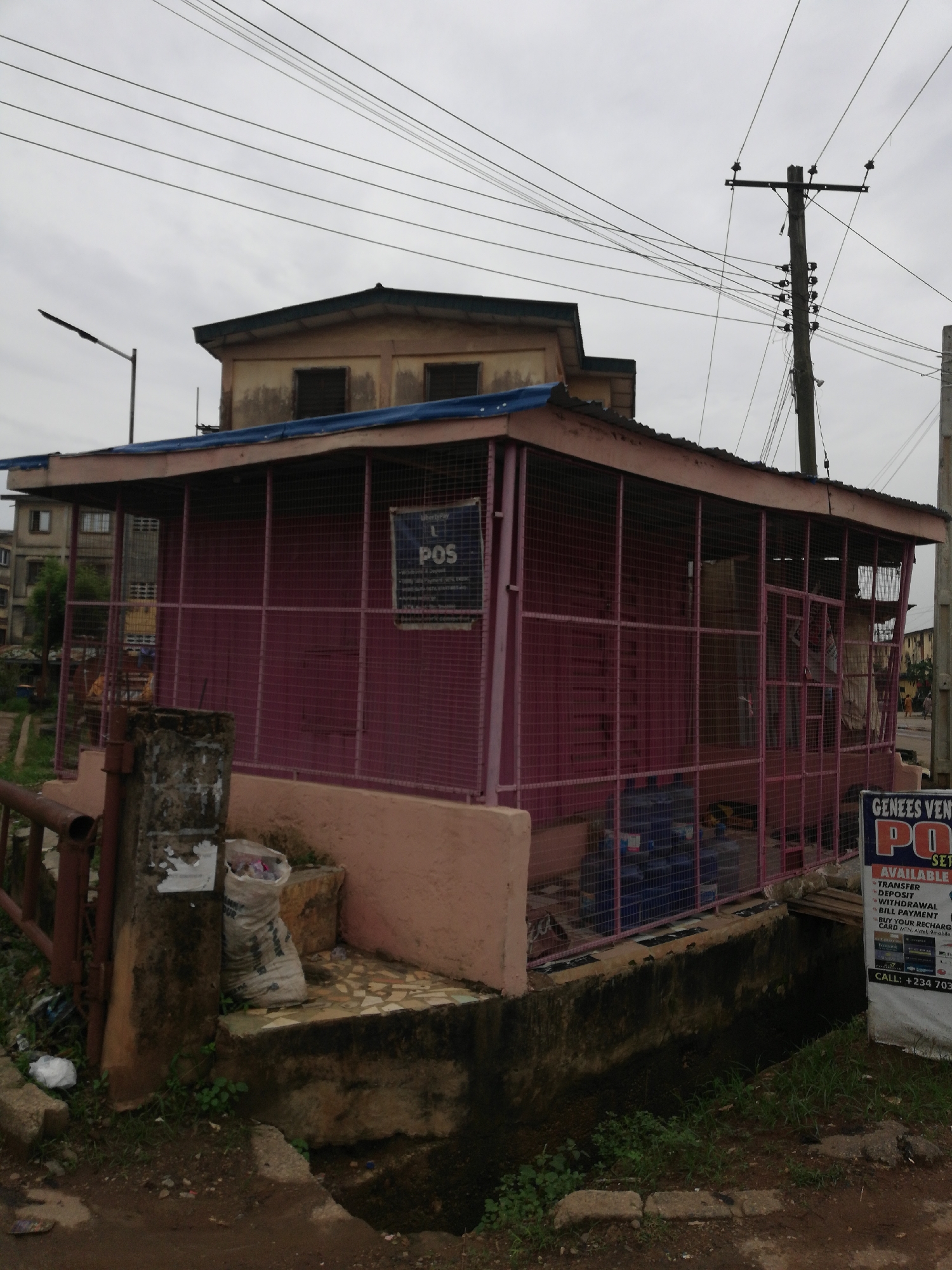 The image shows a small, magenta-colored, caged structure with a blue roof, which appears to be a Point of Sale (POS) business location, likely for financial transactions like money transfers, deposits, withdrawals, and bill payments. A POS advertisement board can be seen to the right of the structure. The structure is situated adjacent to a drainage ditch and a concrete bridge. A residential building stands behind the POS structure. An electrical pole with wires is visible in the background, against a cloudy sky. Debris and litter are scattered around the base of the structure and in the drainage ditch.