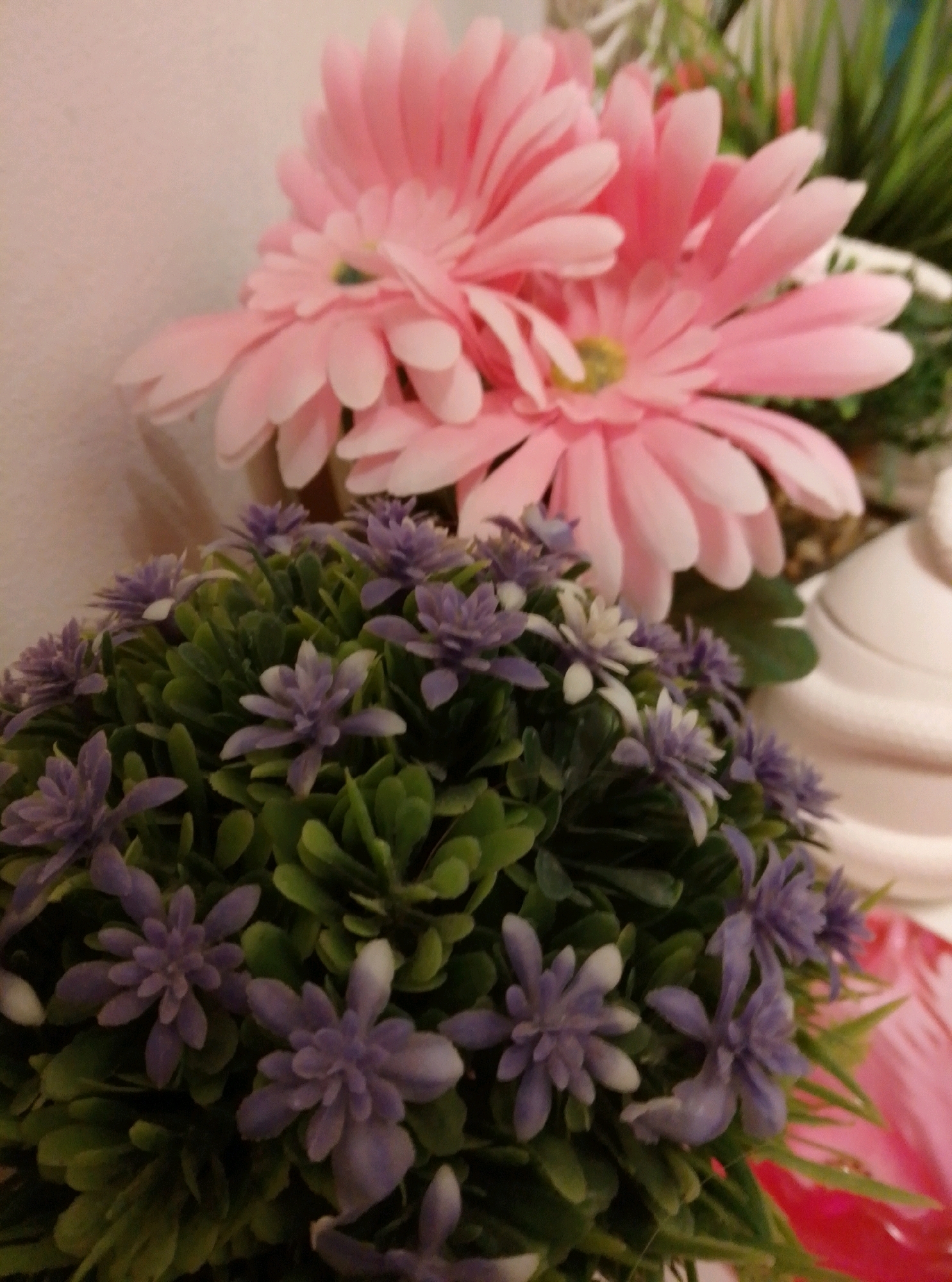 The image displays a close-up view of artificial flowers. In the foreground, a dense arrangement of green foliage is interspersed with small, purple, star-shaped flowers, some of which have white tips. Behind this, two large, pale pink gerbera daisies are visible, their petals fanned out. The background is a plain, light-colored surface, possibly a wall. To the right, a portion of a white ceramic object, possibly a vase or pot, can be seen. The lighting is soft, casting gentle shadows and highlighting the textures of the petals and leaves. The image does not contain any elements that would indicate a specific location in the city of Wedel, Germany.