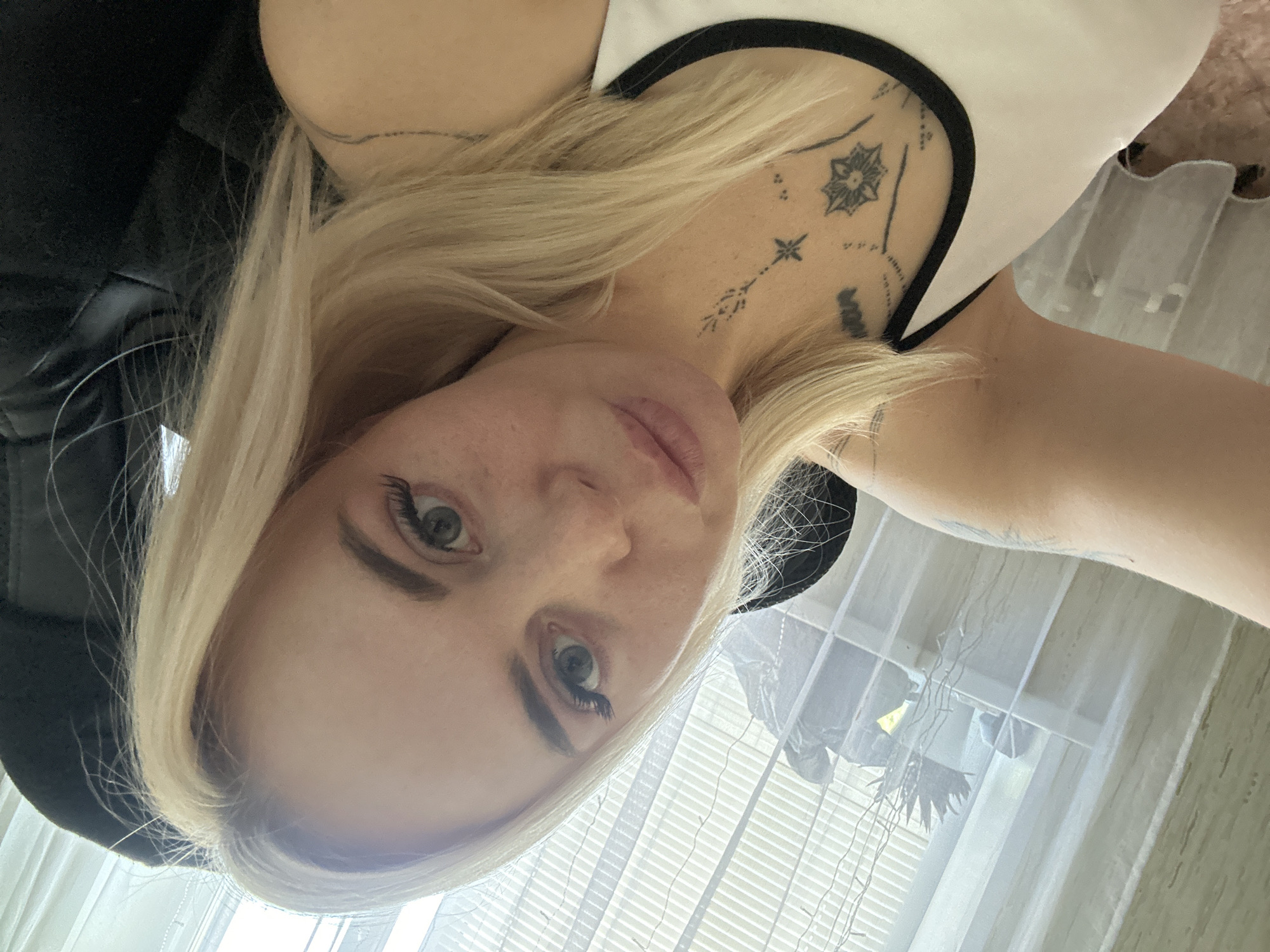 A blonde woman is taking a selfie in an indoor setting. She is wearing a white tank top with black trim. Tattoos are visible on her chest and upper arm. The background shows a window with blinds and sheer curtains, and a black chair. The lighting suggests it is daytime. The exact location within the city is unknown.