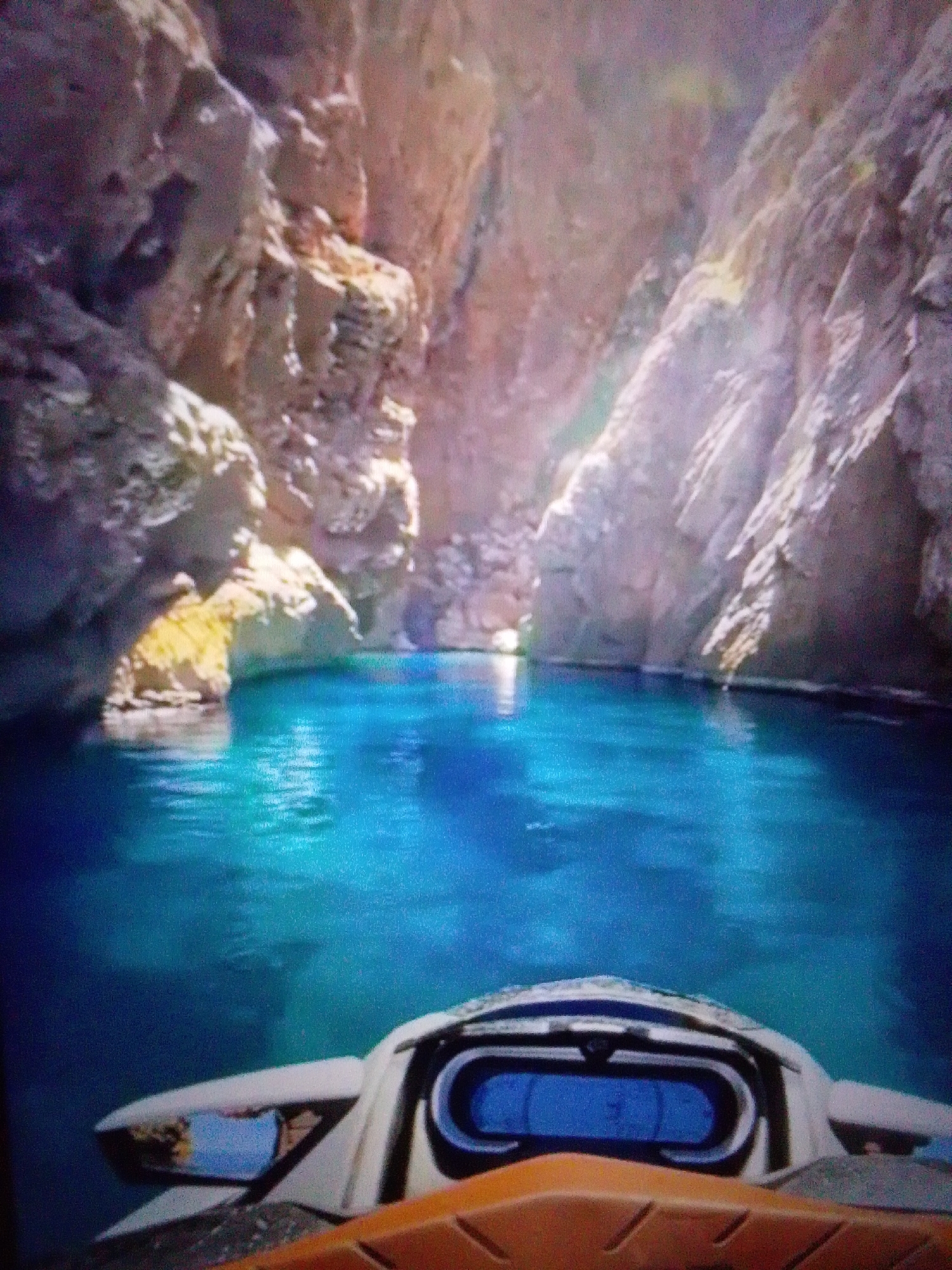 The image shows a first-person perspective of someone riding a jet ski through a narrow canyon with a body of bright blue water. The canyon walls are made of light-colored rock, with some vegetation growing on them. Sunlight streams down into the canyon, illuminating the water and creating reflections on the rock faces. The front of the jet ski is visible in the foreground, including a digital display showing numbers. The scene is calm and serene, with no other people or boats visible. The location is not in the city of Aznakayevo, Russia; it appears to be a natural landscape.