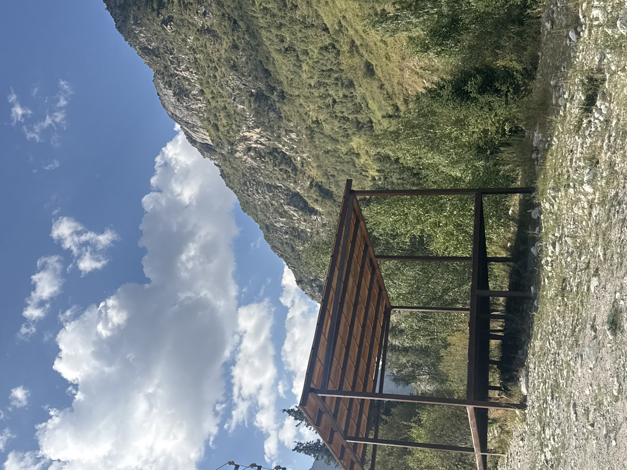 A wooden and metal gazebo stands on a gravelly embankment in front of a lush green mountainside under a bright blue sky dotted with white clouds. The gazebo is raised on stilts and has a flat, slatted wooden roof supported by dark metal beams and posts. The mountain behind it is covered in dense foliage and rock formations. Trees and bushes are visible around the gazebo and on the lower slopes of the mountain. The ground in the foreground is a mixture of rocks, pebbles, and sparse grass. The scene appears to be a scenic outdoor location in Kyrgyzstan.
