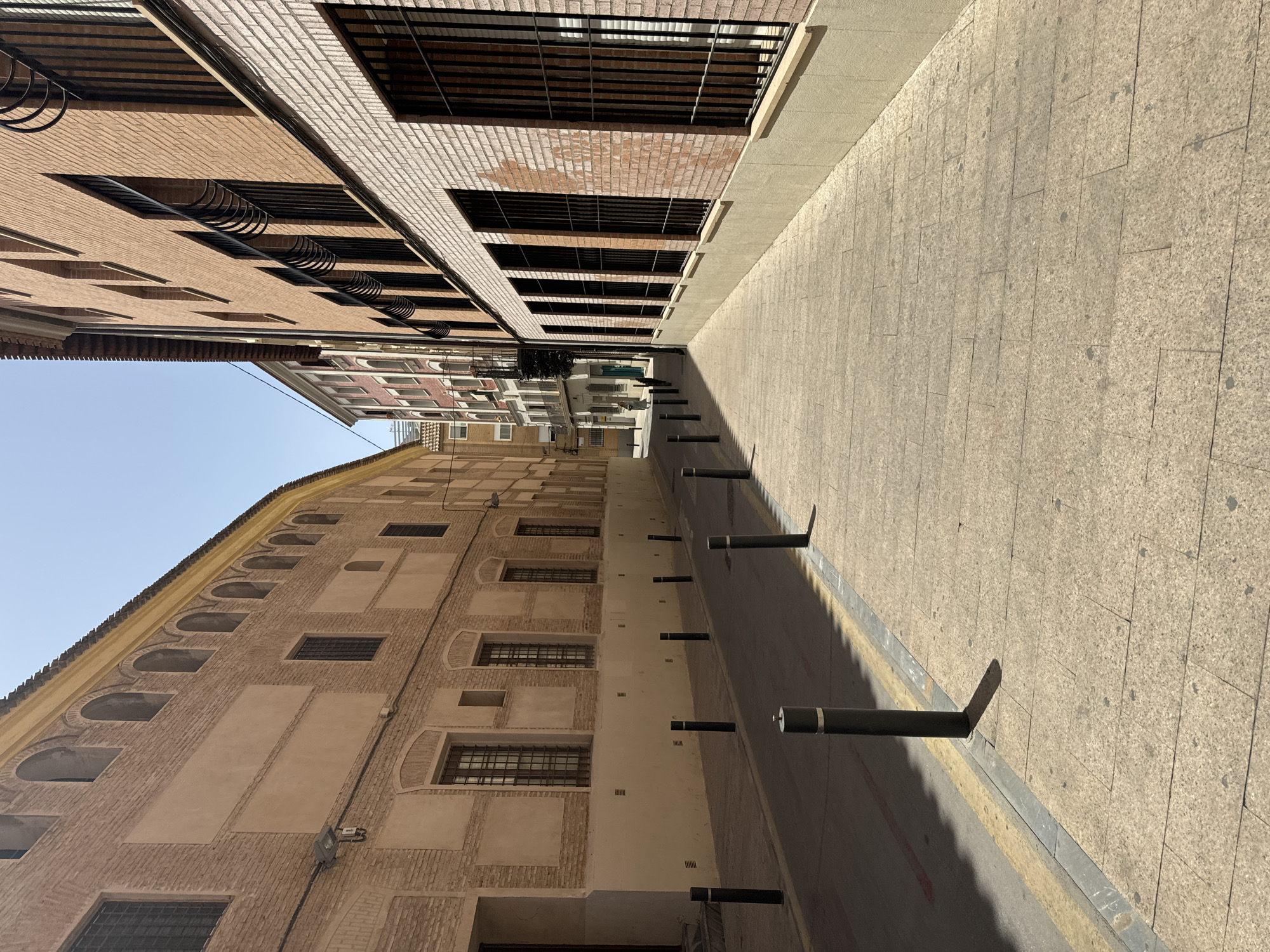 The image depicts a street scene in Murcia, Spain. The perspective is from an elevated viewpoint, looking down a narrow street lined with buildings. On the left, one building features a brick facade with arched windows and smaller, rectangular windows with metal grates. Another building visible further down the street has a lighter-colored brick facade with regularly spaced windows. On the right side of the street, a pedestrian walkway paved with light-colored square tiles is bordered by a darker asphalt road. Along the edge of the walkway, there are several dark, cylindrical bollards spaced at intervals. The sun is casting long shadows from the bollards and buildings onto the walkway, suggesting it is either morning or late afternoon. The sky is clear and blue, indicating fair weather. There are no people visible in the image. The architecture suggests an urban environment with a mix of older and more modern construction.