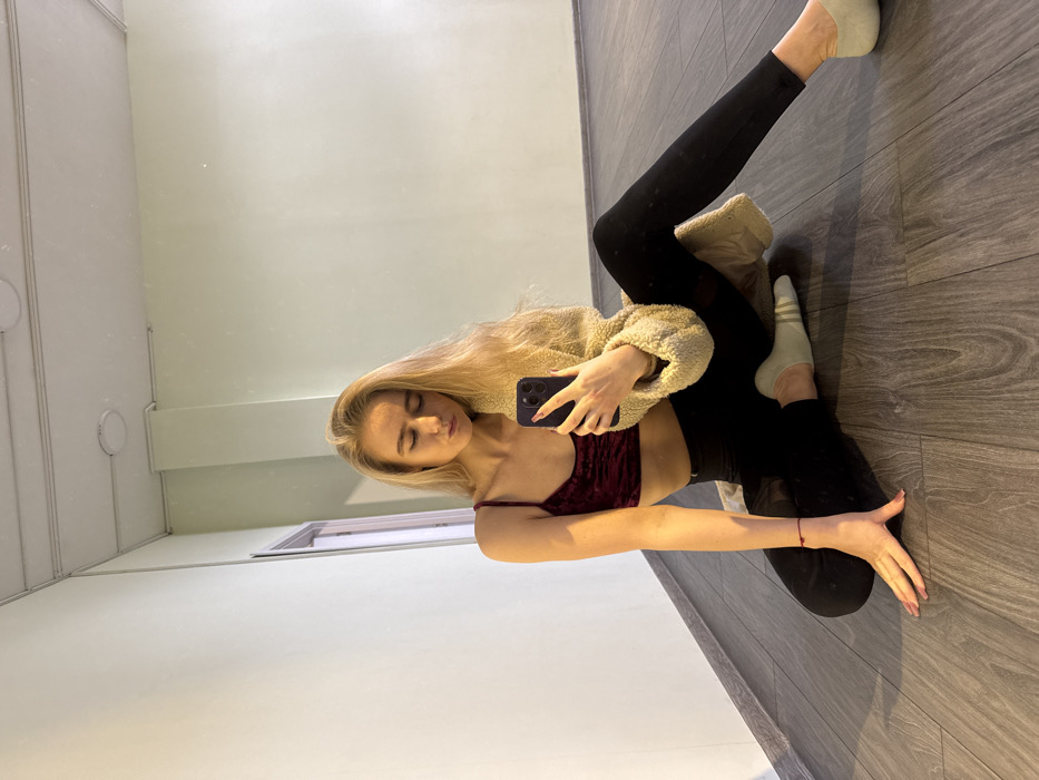 The image displays a young adult female sitting on a dark wood-laminate floor, taking a mirror selfie. She has long blonde hair and is wearing a dark red velvet crop top with thin straps, black leggings, and light green ankle socks. A beige fleecy jacket is draped over her shoulders and back. She holds a dark-colored smartphone in her left hand, positioned for the photo, with her right hand resting flat on the floor. Her eyes are closed or cast downwards, and her lips are pursed. The background consists of plain light green walls. A door frame is partially visible on the left side of the frame, and two circular ceiling lights are reflected in the mirror above. The specific location in the city of Unknown, Unknown, cannot be determined from the visual evidence.
