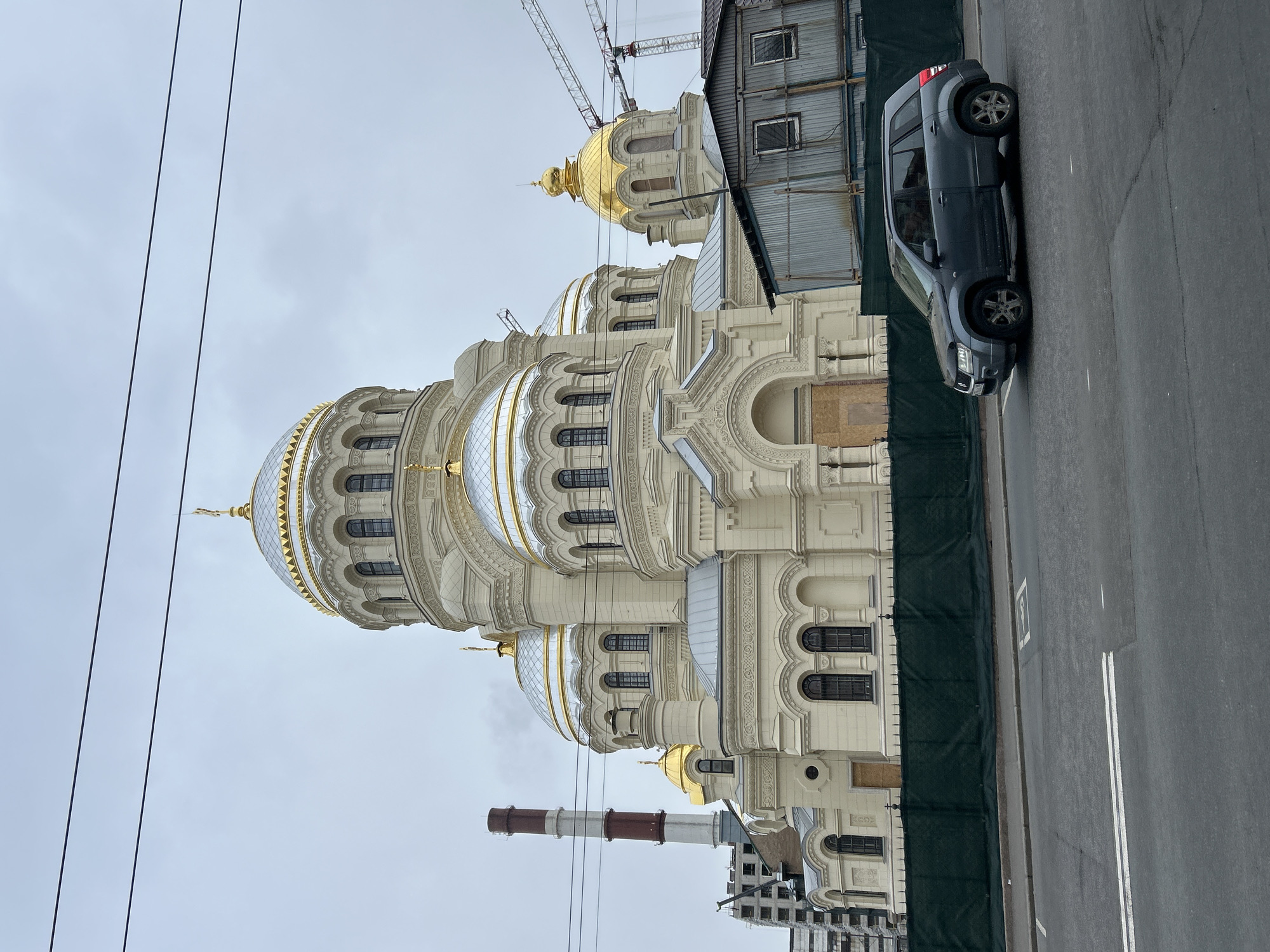 This image, captured in Saint Petersburg, Russia, showcases a grand, ornate cathedral-like building under an overcast sky. The prominent structure is predominantly light cream or beige, characterized by multiple domes; several are large, golden, onion-shaped, while numerous smaller domes are silver with golden detailing, also exhibiting an onion shape. The facade is intricately decorated with rich moldings, pilasters, and arched windows featuring dark frames.
An asphalt street runs parallel to the building on the right, separated by a dark green construction fence. A dark grey SUV, possibly a Suzuki Grand Vitara, is parked along the curb. The road surface displays double white lines and a faded white marking.
In the background and periphery, elements of urban infrastructure and potential construction are visible. A large construction crane extends upwards behind the golden domes on the right, suggesting ongoing building or renovation work. A temporary structure with corrugated metal siding is also present. A tall, red and white striped industrial smokestack rises behind the lower-left portion of the main building. Two thin, dark overhead power lines diagonally traverse the cloudy sky in the upper left.
The scene is set during a cloudy daytime, illuminated by natural light. No people are visible, and there is no discernible text apart from the white painted road markings.