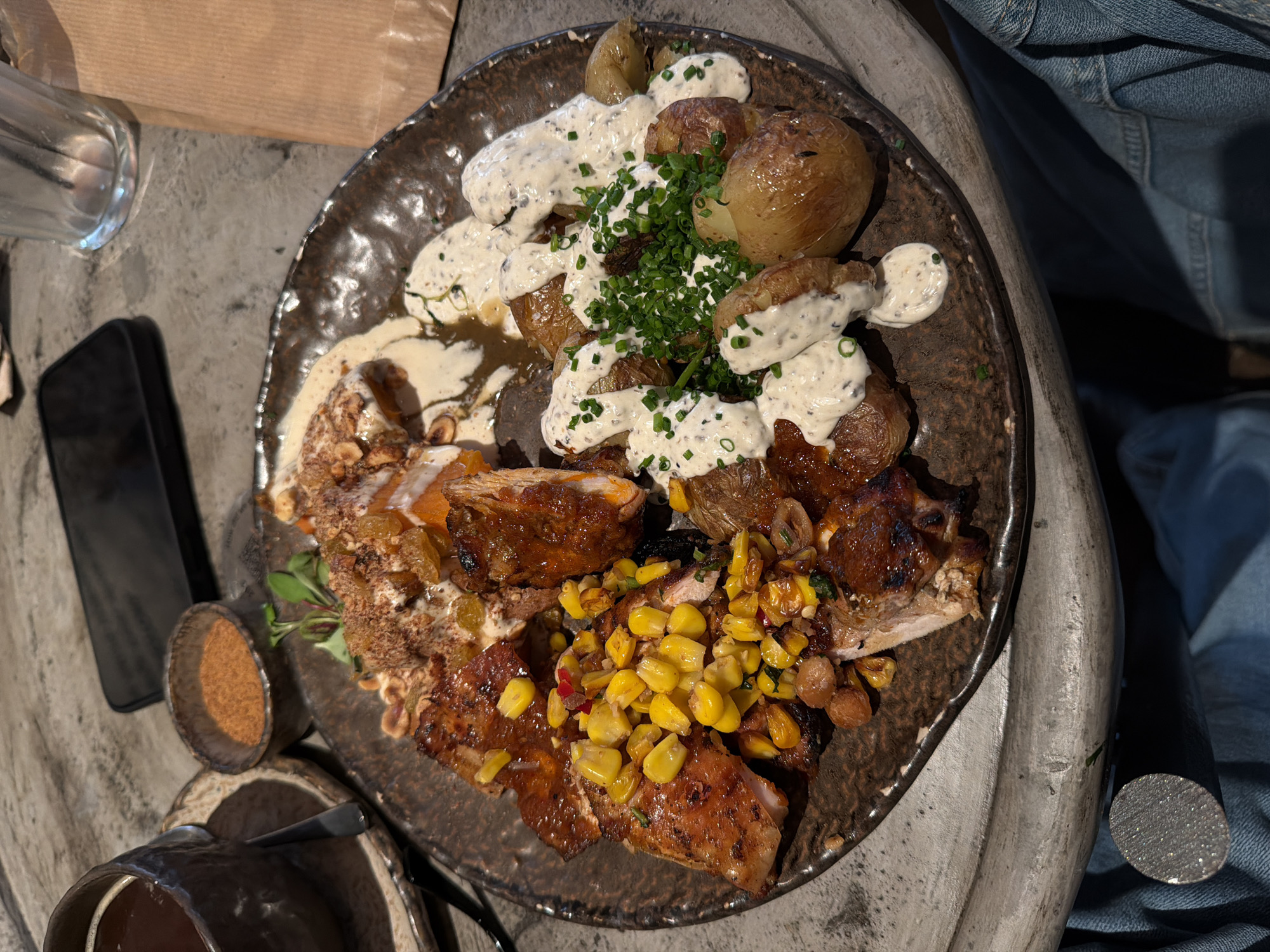 A top-down shot shows a brown ceramic plate filled with food, likely taken in an urban environment in London, United Kingdom. On the plate are roasted potatoes covered in a creamy white sauce and chopped green herbs, alongside grilled meat, possibly chicken, glazed with a dark sauce. There is also a mix of corn kernels and beans. To the left of the plate, there is a smartphone, a small cup with a brownish powder, and a cup with a dark liquid. The plate sits on a stone-like surface next to someone wearing denim.