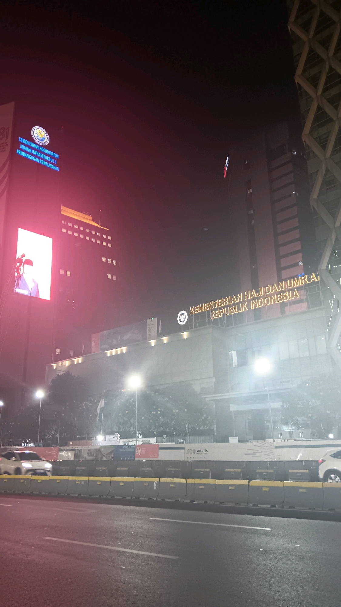 The image depicts a nighttime view of a government building in Central Jakarta, Indonesia. The building, illuminated by prominent neon signs, displays the text "KEMENTERIAN KOORDINATOR BIDANG INFRASTRUKTUR & PEMBANGUNAN KEWILAYAHAN" and "KEMENTERIAN HAJI DAN UMRA: REPUBLIK INDONESIA". A large digital screen on the left side of the building shows a person in a red frame. The scene is set along a road with traffic barriers and a passing car. The overall atmosphere is urban and illuminated by artificial lights, with some atmospheric haze present.