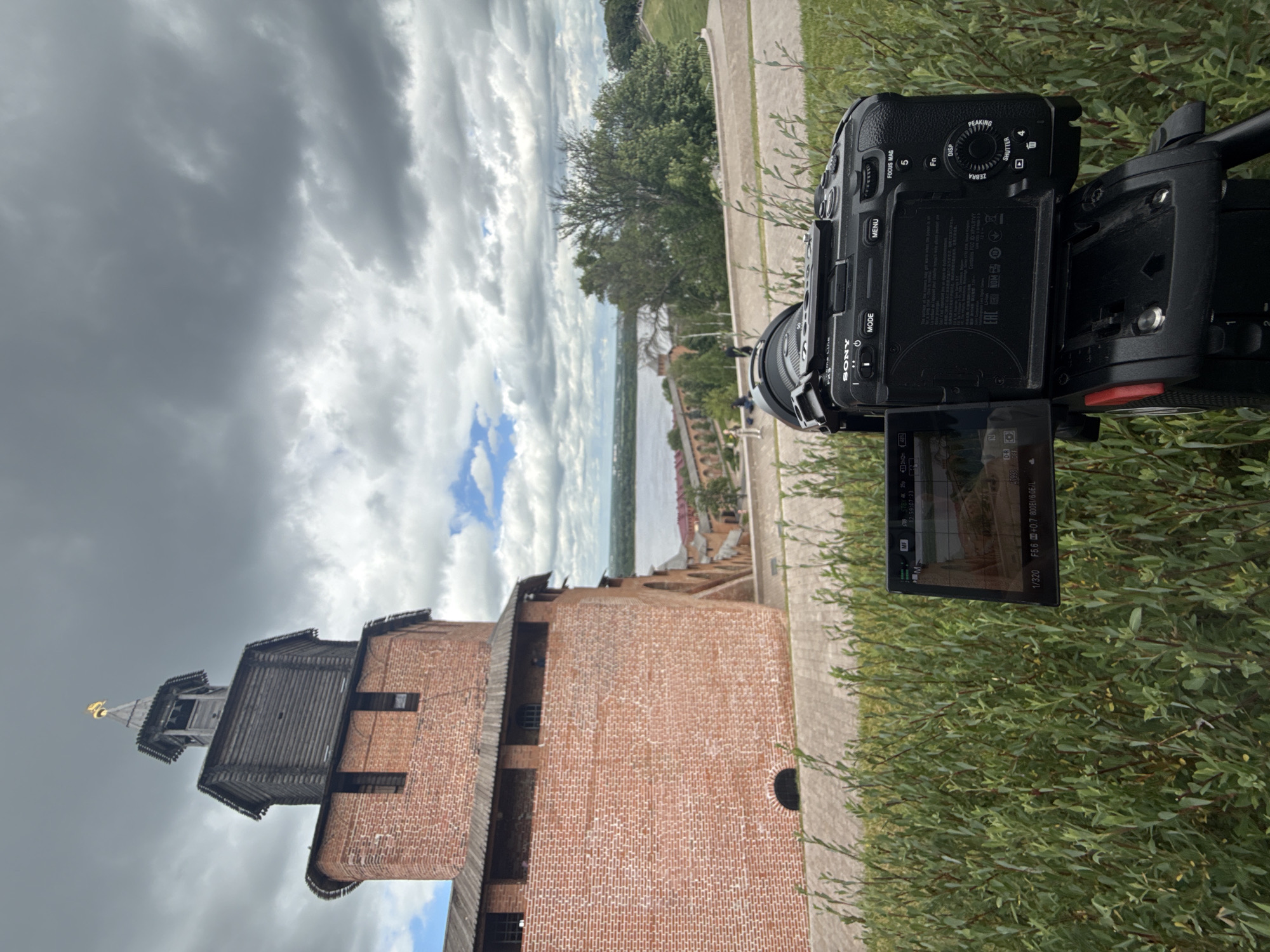 The image shows a Sony camera on a tripod, positioned in front of a brick tower, likely part of the Nizhny Novgorod Kremlin, overlooking the Volga River. The tower is made of red brick with a wooden top section and a golden spire. The sky is cloudy, with some patches of blue showing through. In the background, the river is visible, along with trees and a distant cityscape. The camera's screen displays a view of the tower and the surrounding landscape. There are a few blurred figures in the distance, but their actions are not discernible. The foreground consists of green bushes.