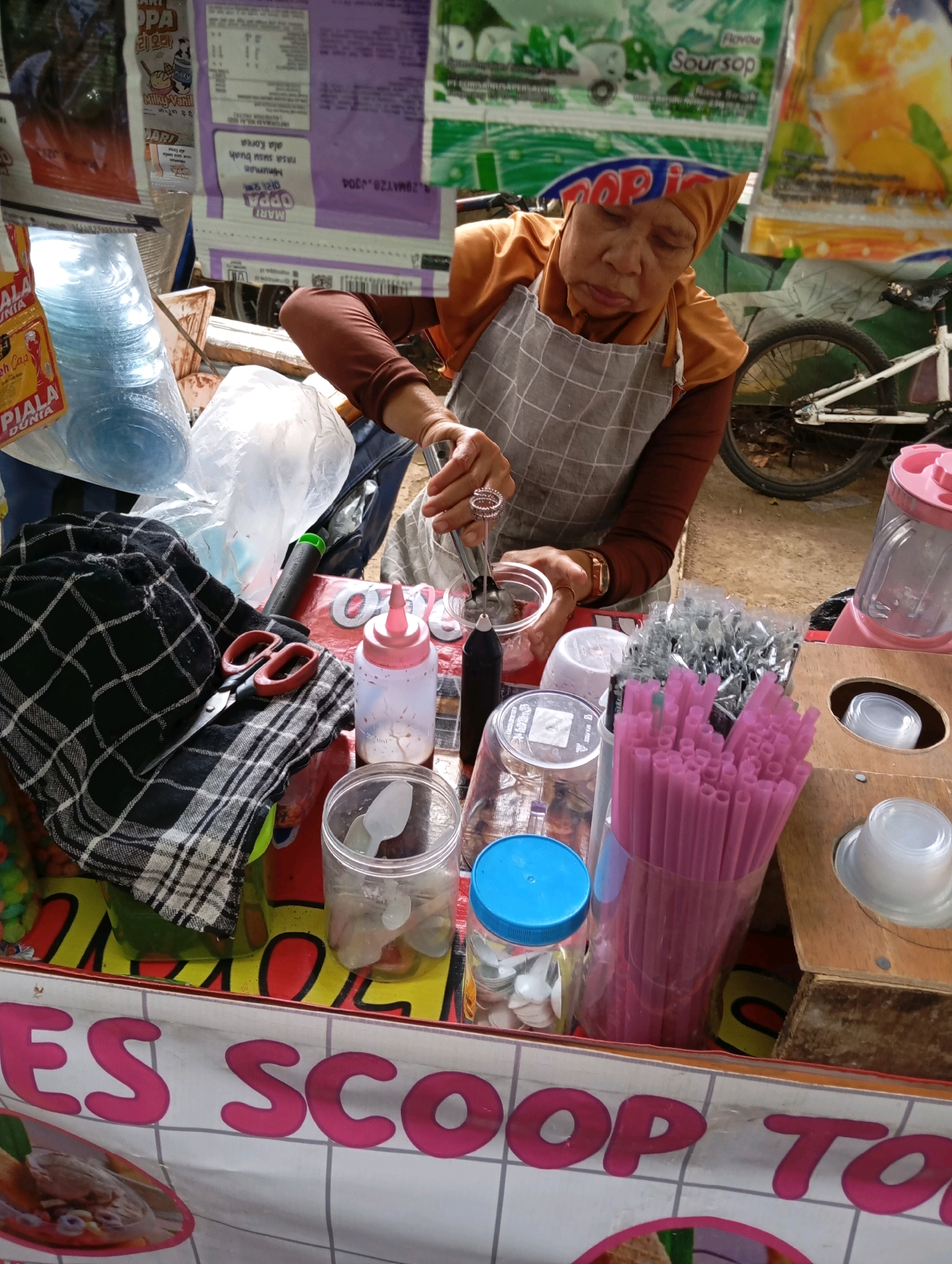 The image depicts a street vendor in Bandung City, Indonesia, preparing drinks at her cart. The vendor, an adult woman wearing a brown headscarf and an apron, is actively engaged in her work, holding a tool that appears to be an immersion blender to mix ingredients in a clear plastic cup. Surrounding her are various items essential for her business, including stacks of disposable plastic cups, pink drinking straws, a pink squeeze bottle, and other containers filled with what seem to be ingredients or utensils. In the background, a bicycle is visible, suggesting a typical urban street setting. The cart itself is adorned with product packaging and a banner displaying the words "ES SCOOP TA". The scene conveys the everyday hustle of a street food vendor in a bustling Indonesian city.