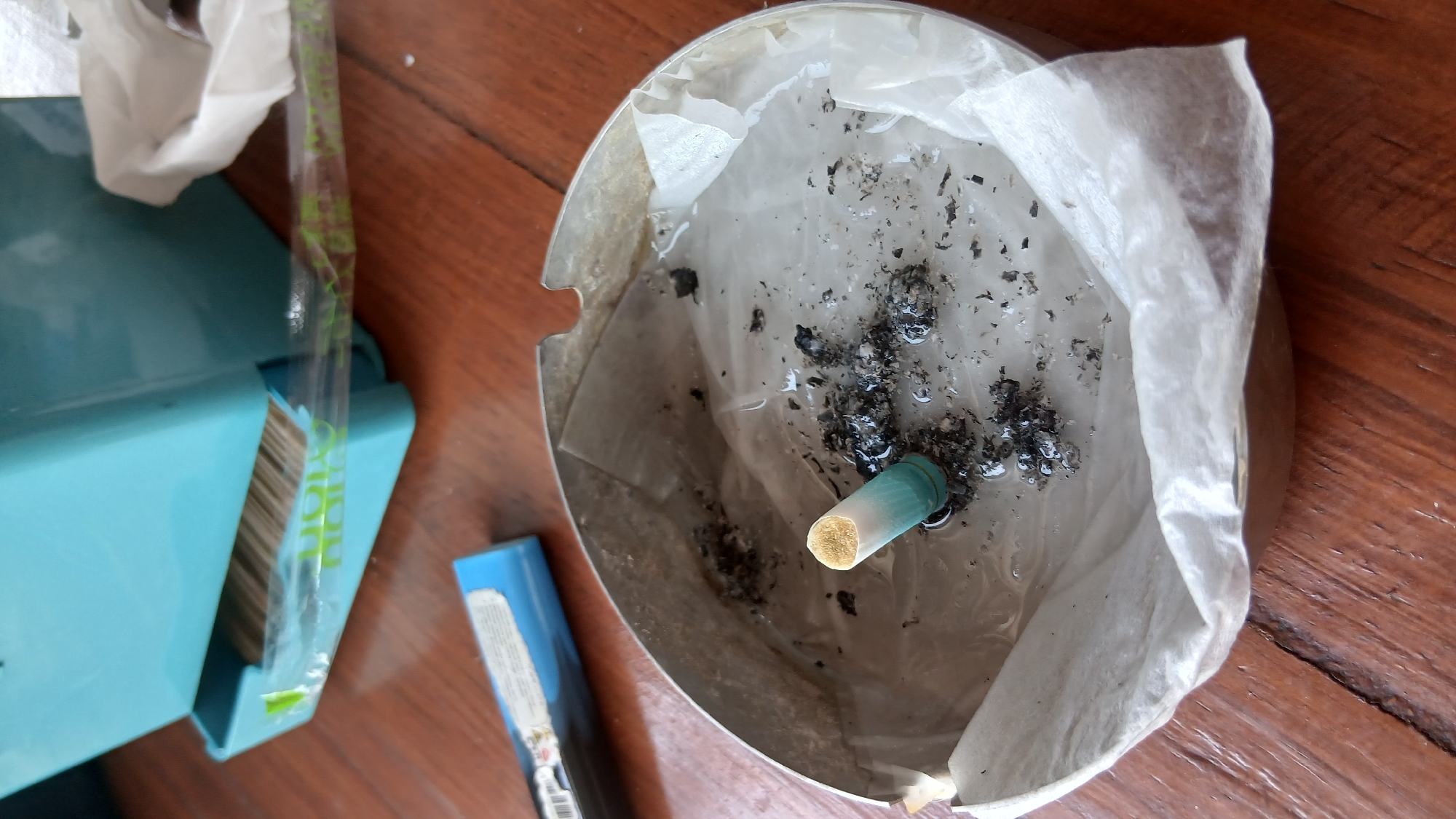 The image depicts an ashtray on a wooden table. The ashtray is round and metallic, lined with white tissue paper. It contains several cigarette butts, ashes, and a small amount of liquid, suggesting it has been recently used. In the foreground, a single cigarette butt stands upright, its filter end visible and partially submerged in the liquid. Behind the ashtray, a blue lighter lies on the table. Further in the background, a teal plastic dispenser filled with toothpicks is visible. The scene appears to be an indoor setting. The image does not contain information that would allow for the determination of its location in Banda Aceh, Indonesia.