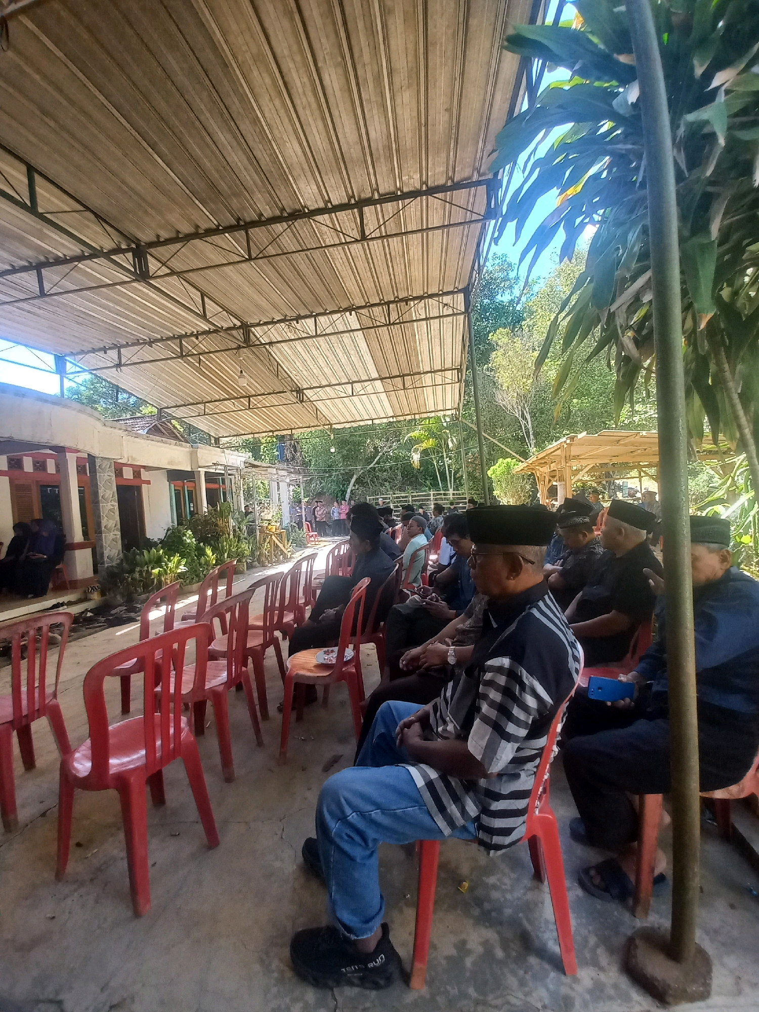 The image depicts an outdoor gathering, possibly a ceremony or meeting, under a corrugated metal roof supported by metal beams. Many people, predominantly men, are seated on red plastic chairs arranged in rows. Most of the men are wearing traditional Indonesian attire, including peci (a type of cap). They are facing towards the left of the frame, suggesting a focal point or speaker in that direction, though not clearly visible. The environment appears to be semi-rural, with lush green vegetation visible in the background and a building with a traditional Indonesian architectural style on the left. The ground is paved with concrete. One individual in the foreground is wearing a striped shirt and blue jeans, holding a blue smartphone. The overall atmosphere seems formal and communal. The specific location in Sukoharjo, Indonesia, cannot be determined from the visual information provided in the image.