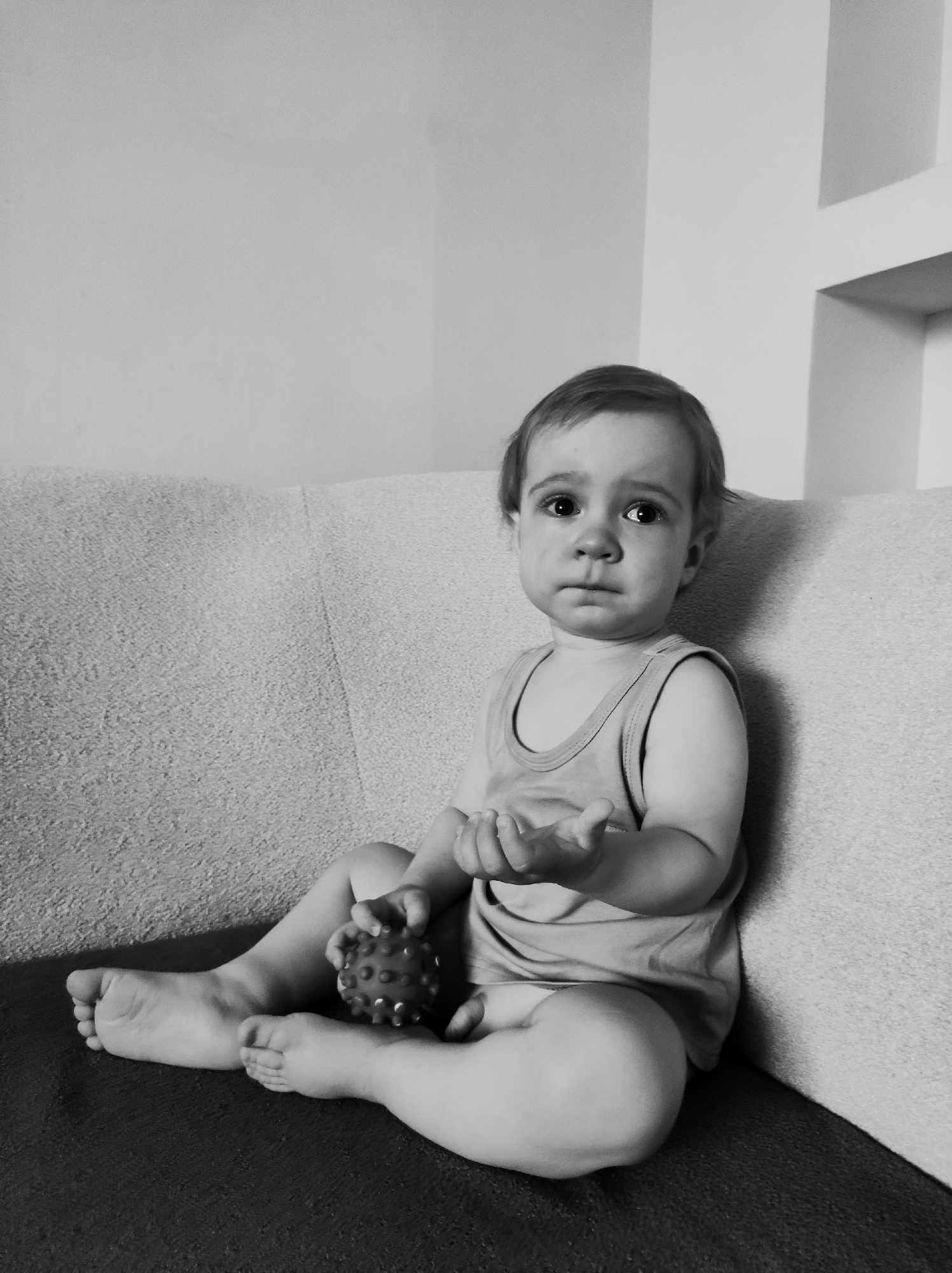 A black and white photograph depicts a toddler seated on a sofa. The child is wearing a tank top and is barefoot. The toddler is holding a spiky ball in one hand, while the other is slightly outstretched. The background includes a plain wall and a recessed section of the wall. There is no indication this picture was taken in the city of Фоки, Russia.