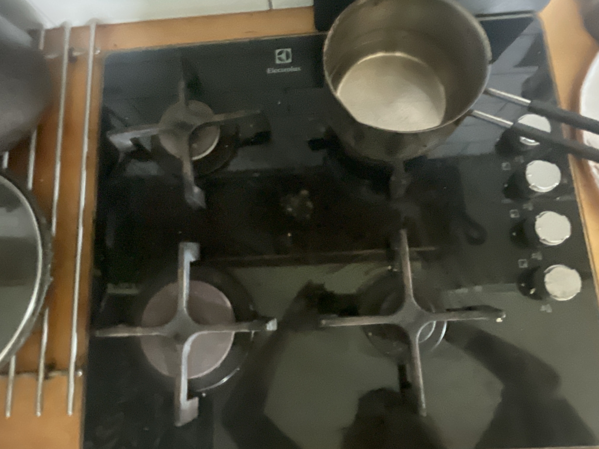 The image shows a close-up, overhead view of a black gas stovetop in a kitchen. The stovetop has four burners, with two burners visible and partially obscured by cookware. A small, metal saucepan with a light-colored interior is positioned on the right side of the stovetop. The stovetop has control knobs at the bottom. Above the stovetop, there are metal racks, and the edge of a large pot or pan is visible at the top of the image. The overall environment appears to be domestic. The provided context about the location being in Мухавецкі сельскі Савет, Belarus, is not visually discernible from the image itself.