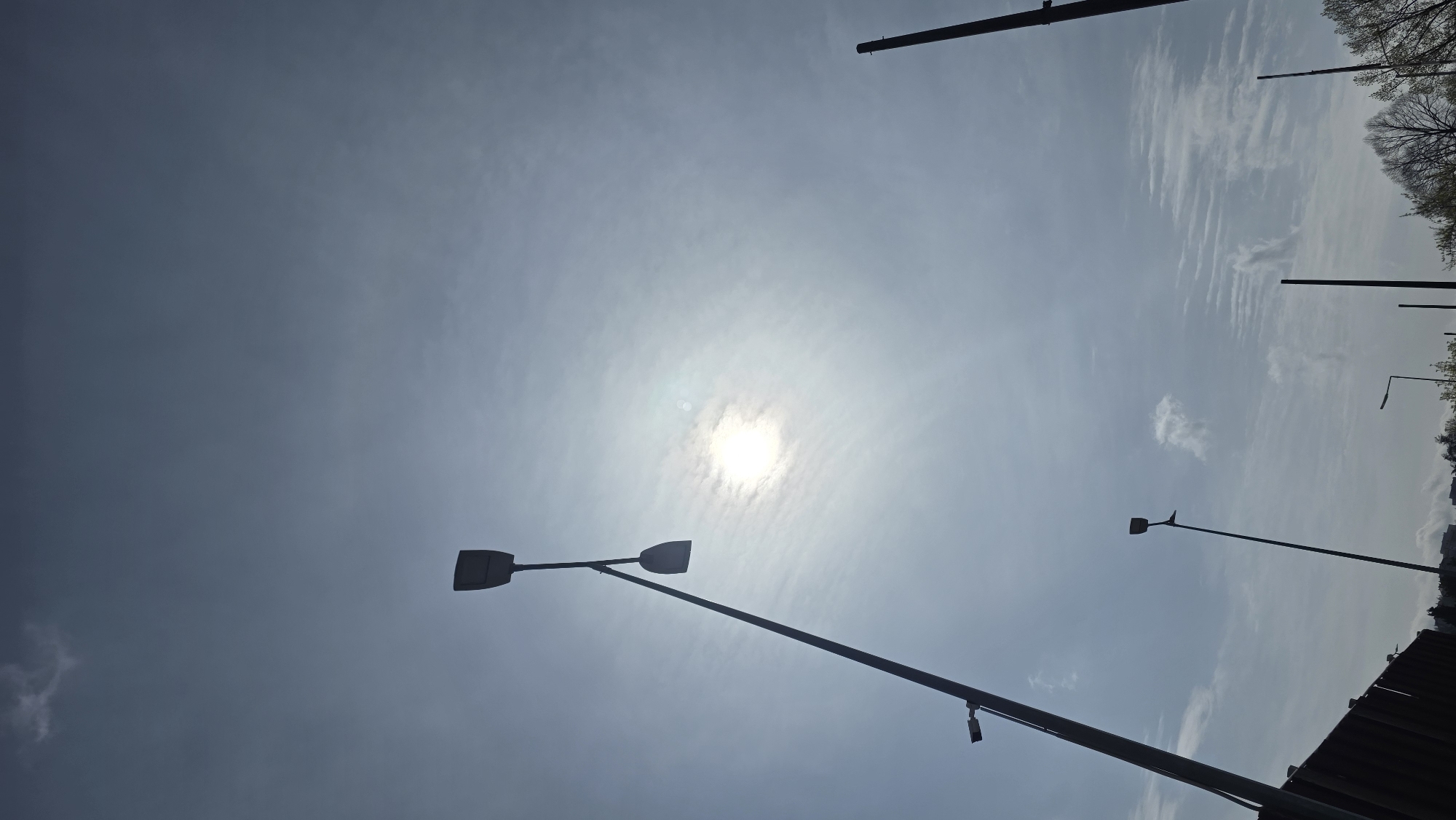 The image shows a sky with the sun visible, partially obscured by clouds, in Blagoevgrad, Bulgaria. There are several streetlights visible, some with two lamps and others with one. There are also vertical poles and some trees in the background. A structure with a dark, possibly metal or concrete edge is visible on the lower right.