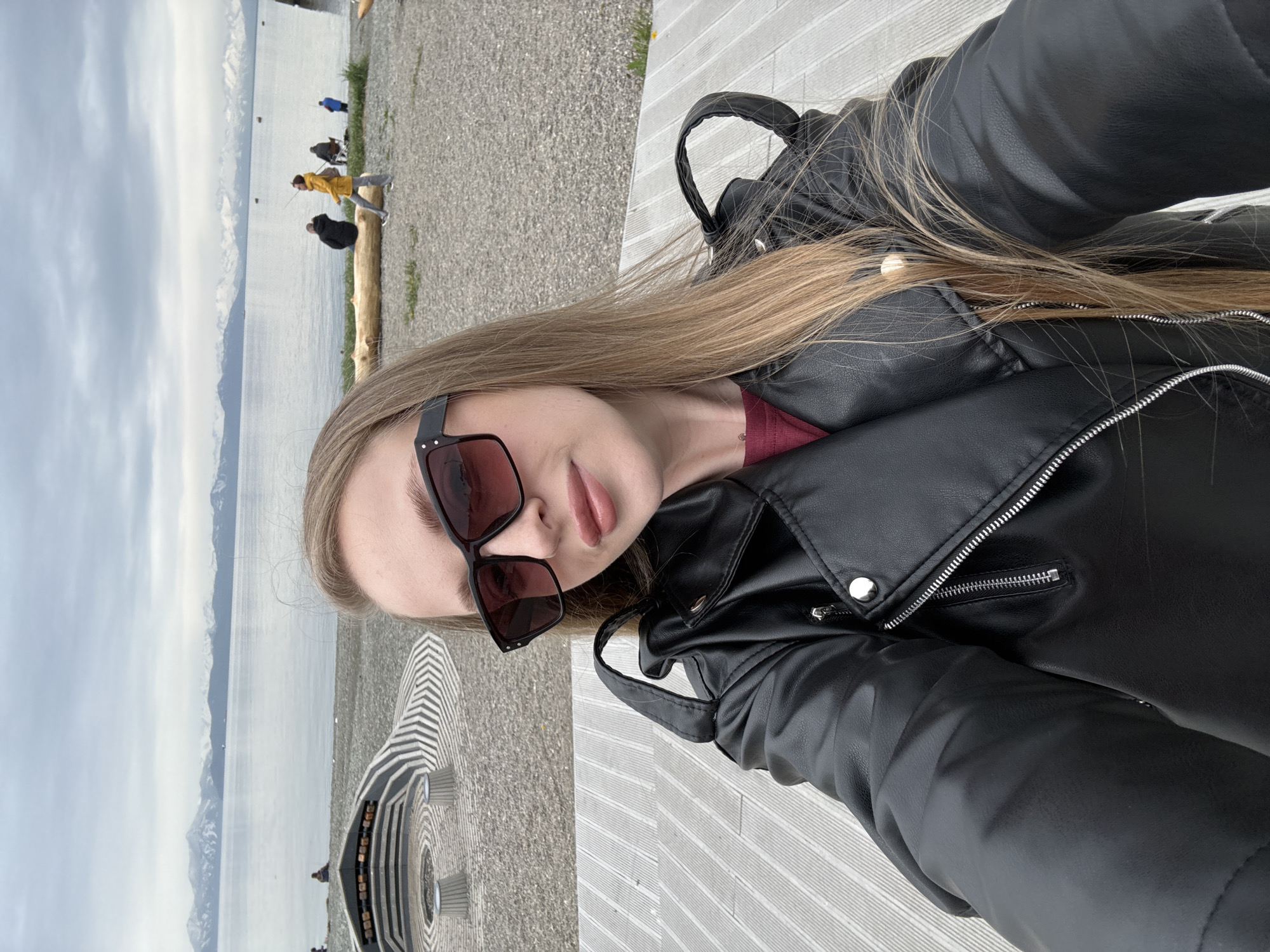 A young Caucasian woman with long blonde hair and sunglasses is taking a selfie outdoors. She is wearing a black leather jacket over a maroon shirt. The background shows a body of water with a mountain range in the distance. The foreground features a wooden deck. Other people are visible in the background, some sitting on the shore and others fishing. The overall setting suggests a coastal area, possibly Avacha Bay in Petropavlovsk-Kamchatsky, Russia, given the mountainous coastline. The sky is overcast.