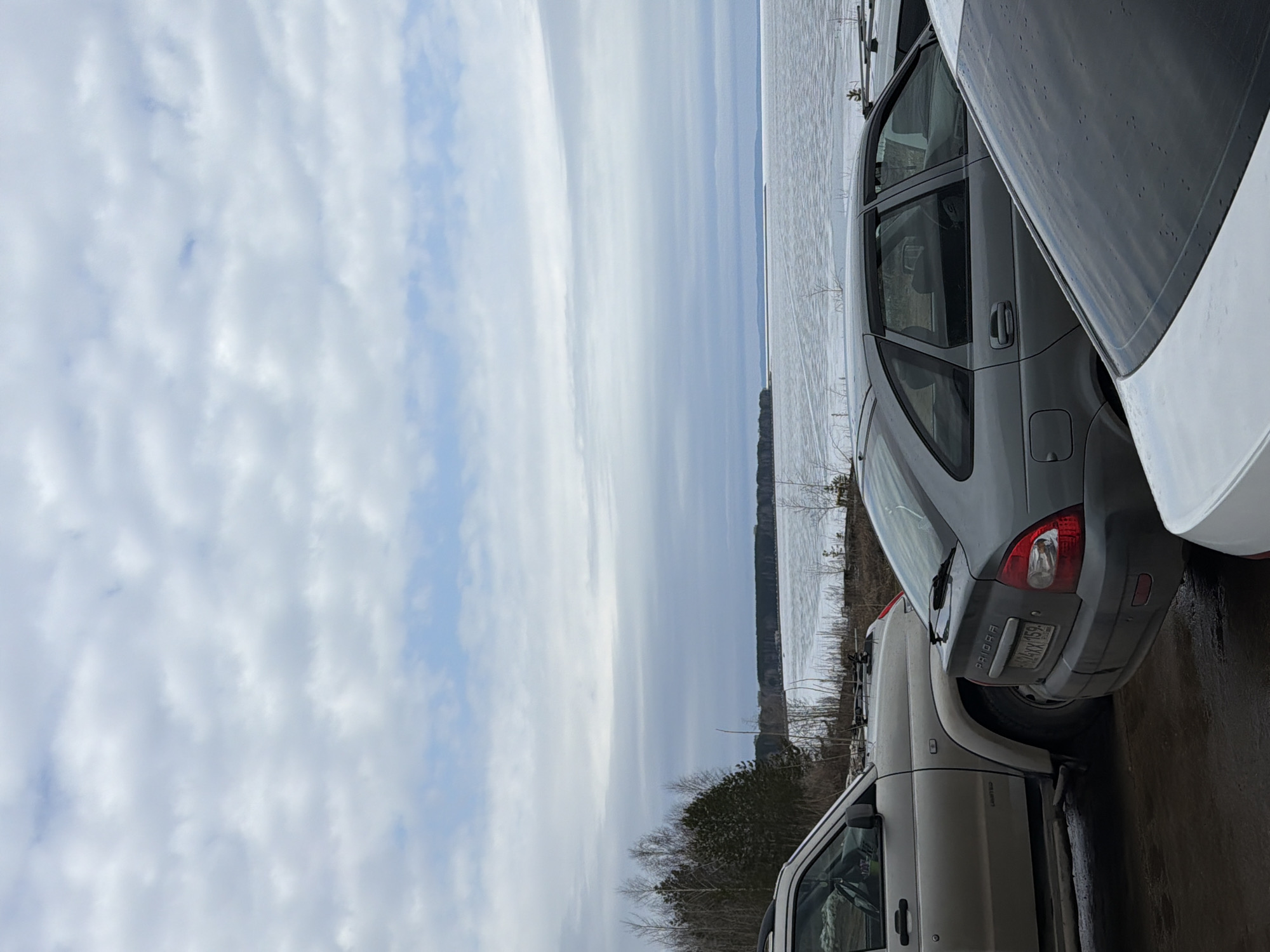 The image shows a scene in Chaikovsky, Russia, featuring a cloudy sky and a partially frozen body of water. A forested area is visible along the shoreline. Three parked vehicles are in the foreground. The registration plate of the middle vehicle reads "A XXX 159".