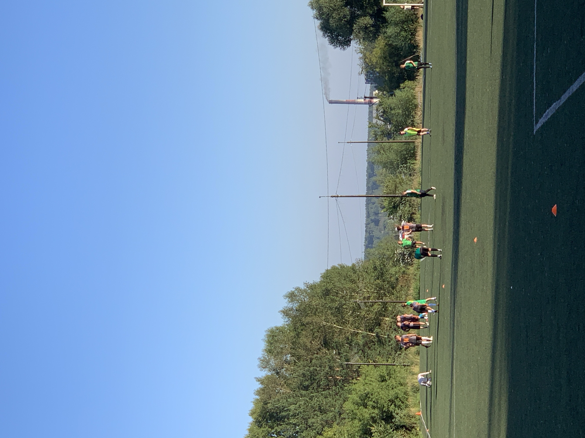An outdoor daytime scene shows a group of young men in athletic attire on a green turf field, likely a soccer field. The men are engaged in what appears to be a practice session, with some gathered in small groups and others scattered across the field. One individual is on the ground, possibly in the process of stretching or recovering. The field is marked with white lines, indicating boundaries and sections of play. Orange cones are strategically placed on the field, suggesting drills or training exercises.
In the background, a line of trees borders the field, with a power line tower visible further back. Beyond the trees and power lines, a factory smokestack with visible smoke billowing from its top is prominent in the distance, against a clear blue sky. The overall impression is that of an industrial or semi-industrial area. Based on the context, the location is likely the city of Prokopyevsk, Russia, given the presence of similar industrial elements. The lighting suggests it is either early morning or late afternoon.
