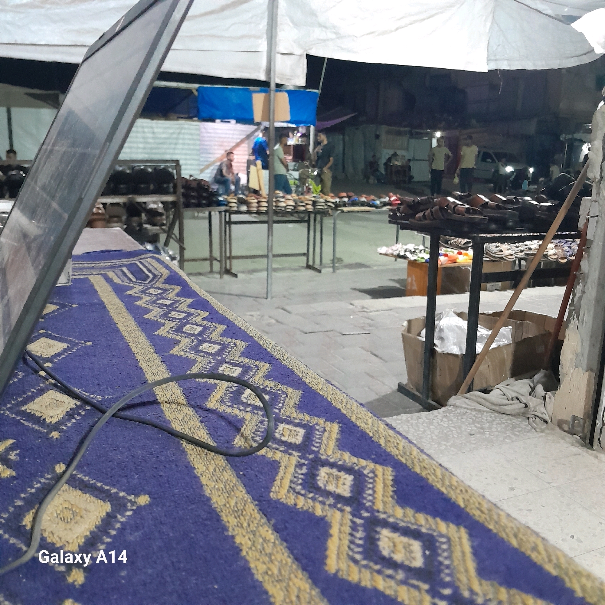The image depicts an outdoor market scene, likely in Khan Yunis, Palestinian Territories. In the foreground, a blue and yellow patterned rug is visible, with a black cable running across it. A portion of a reflective screen, possibly a monitor or tablet, is angled into the frame from the left.
The background reveals a market with stalls displaying various goods, primarily footwear. Several metal stands hold numerous pairs of sandals and flip-flops. In the mid-ground, a few individuals are present, some appearing to be vendors or shoppers. One person in a blue shirt is standing near a stall, and another in a light green shirt is engaged in conversation with a man wearing a dark shirt and khaki pants. Further back, more stalls and structures are visible, along with parked vehicles and a larger tent-like canopy. The overall impression is of a bustling commercial area, possibly operating in the evening or at night, given the artificial lighting.