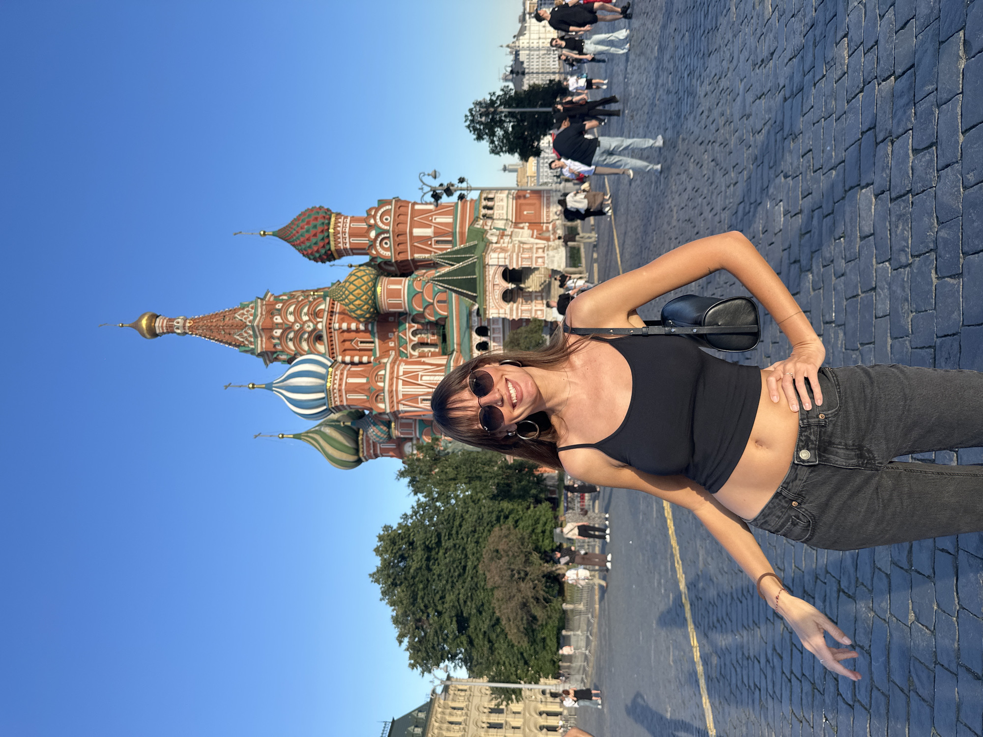 A woman stands in Red Square, Moscow, in front of St. Basil's Cathedral. She is wearing a black tank top, dark wash jeans, sunglasses, and large hoop earrings. A black purse is slung over her shoulder. She is smiling and has her hands on her hips. The ground is paved with cobblestones. Several other people are visible in the background, walking around the square. The sky is clear and blue.