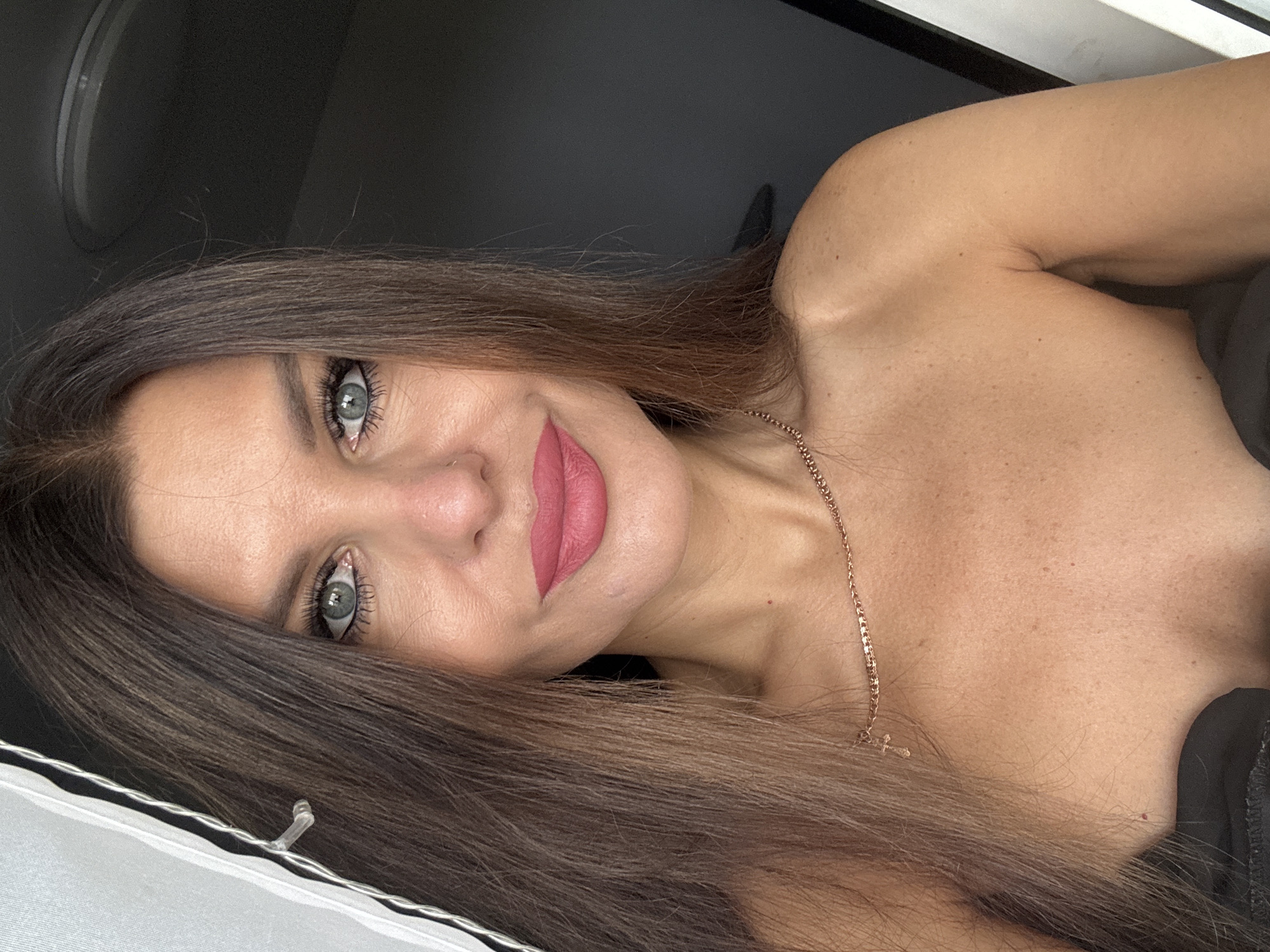 A woman is pictured from the shoulders up. She has long, brown hair that falls to her shoulders. Her eyes are blue and she is wearing pink lipstick. She is wearing a gold chain necklace with a cross pendant. The background is dark, with a white curtain on the left side of the image. The location is Chaikovsky, Russia.