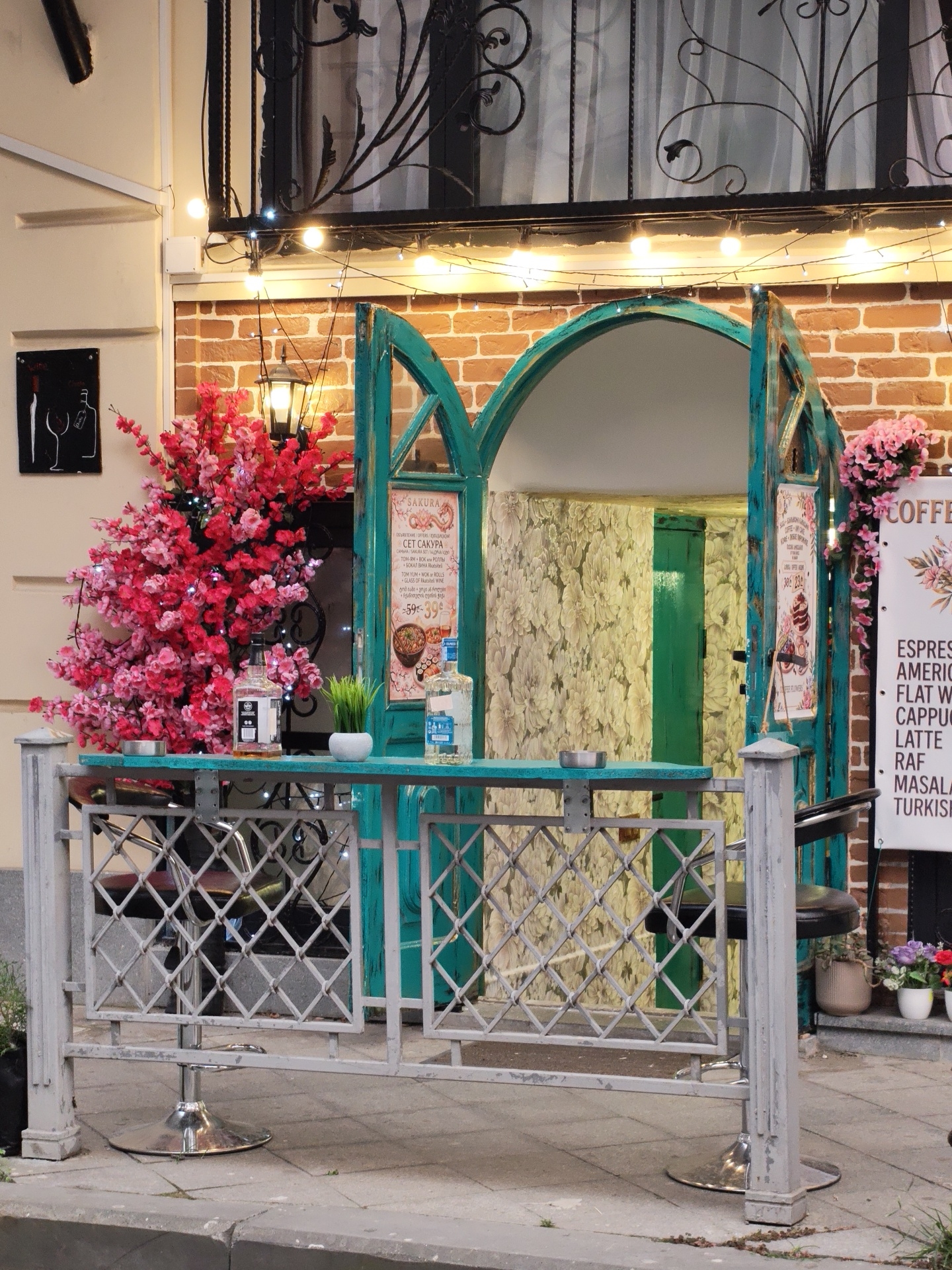 The image depicts a street-level view of a cafe's outdoor seating area in Batumi, Georgia. The cafe entrance is framed by ornate teal archways, leading into a space with floral wallpaper. A turquoise counter displays two bottles of clear liquid, one with a black label and the other with a white label, and a small potted plant. Behind the counter, a neon lamp hangs, illuminating a decorative tree with pink and red blossoms. The cafe is decorated with string lights. A menu board is visible to the right of the entrance, listing various coffee drinks. The outdoor seating area is cordoned off by a grey metal fence with a diamond pattern. A bar stool with a black seat is visible on the right. The cafe is situated on a sidewalk in front of a brick building.