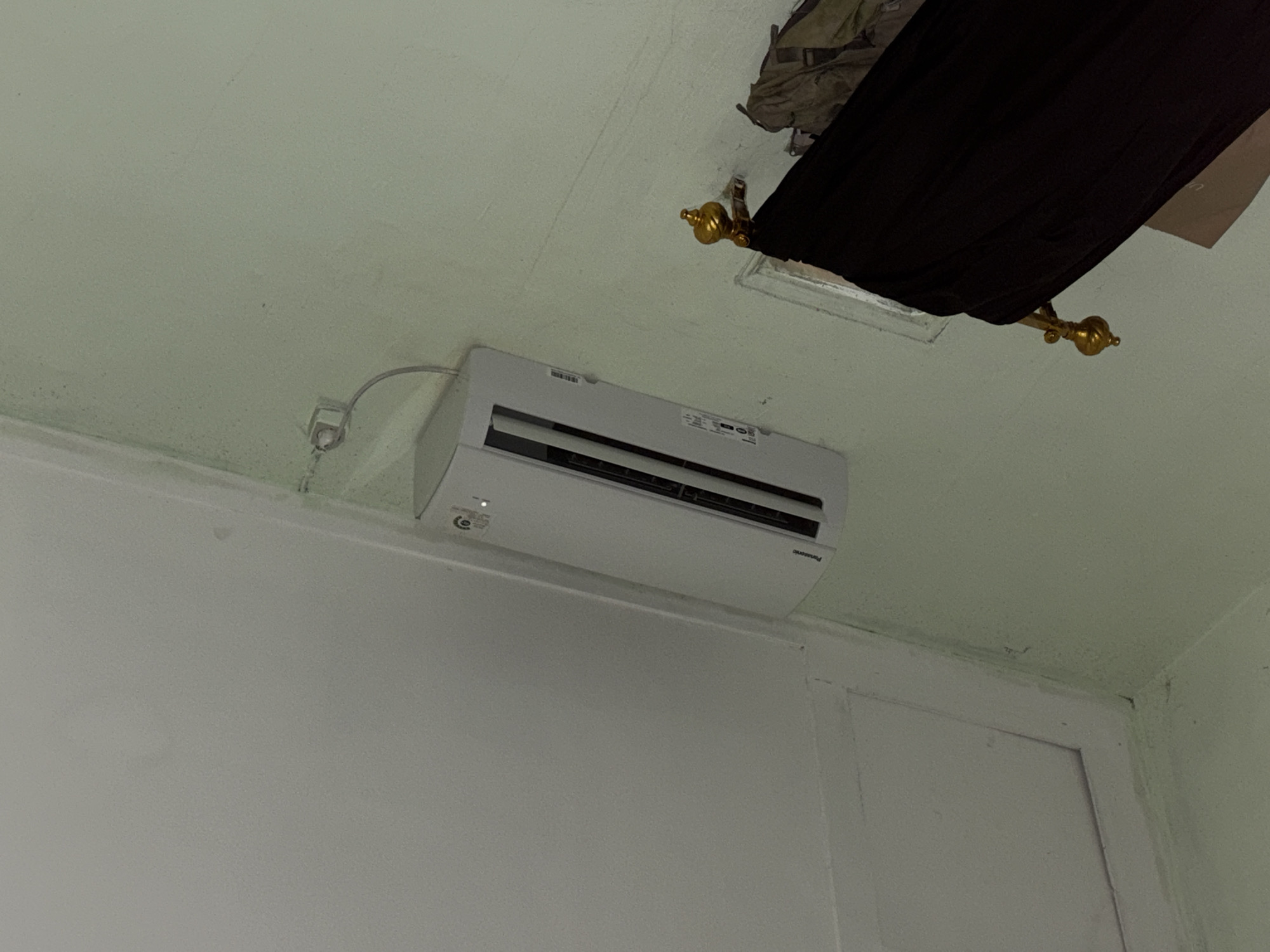 In a room, an indoor air conditioning unit is mounted high on a pale green wall. A white wire runs from the unit to a wall outlet. To the right of the air conditioner, a decorative gold finial is attached to a dark object, possibly part of a piece of furniture or a canopy. The wall appears to have some discoloration or mold in the upper right corner. The ceiling is white and shows a raised rectangular section. The overall scene suggests an indoor residential setting. The location is identified as Tupah, Indonesia.